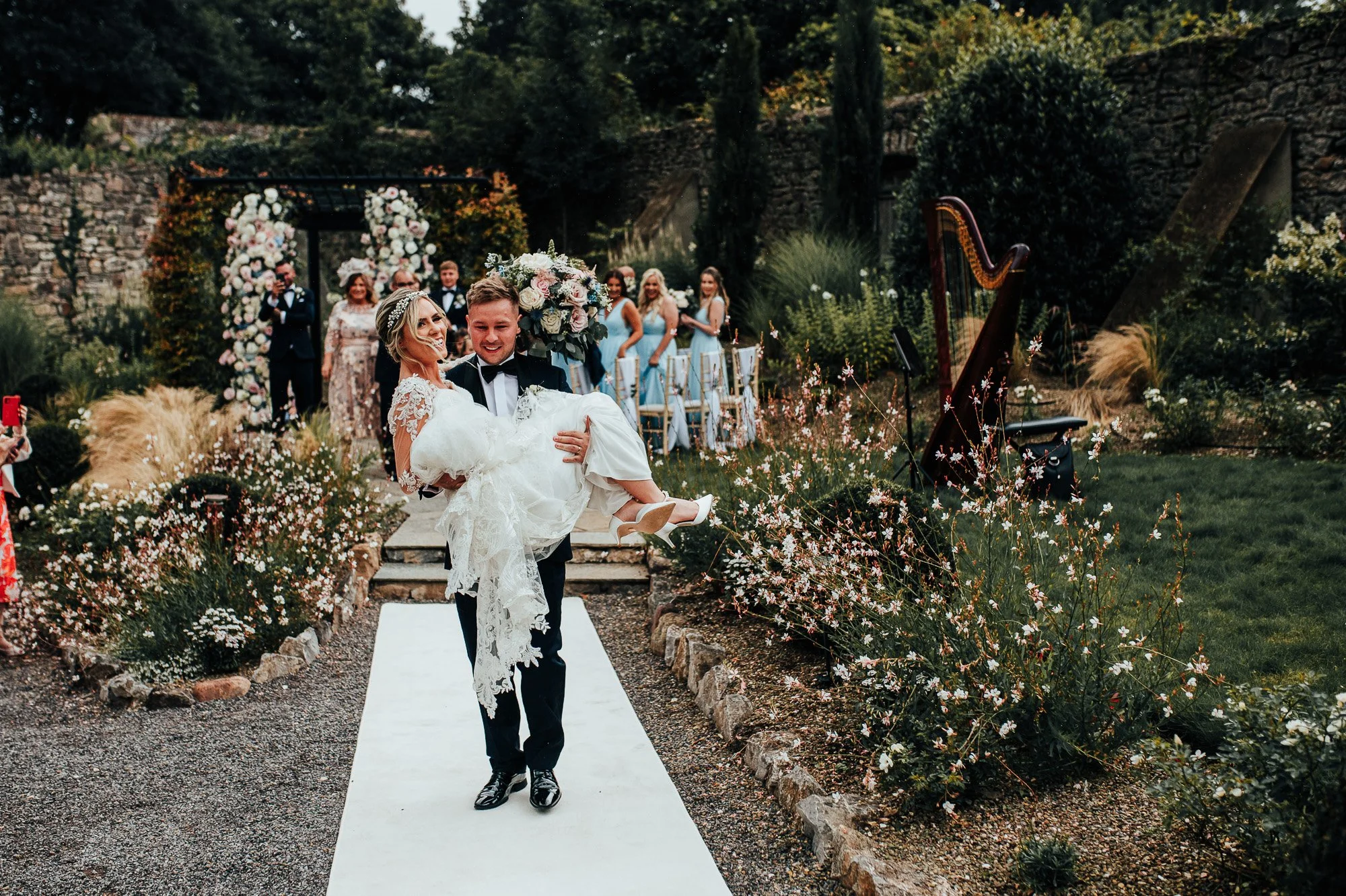 Bride and groom in wedding attire, groom carrying bride down garden path, surrounded by floral decorations, harp in background, wedding party in blue dresses, outdoor wedding setting.