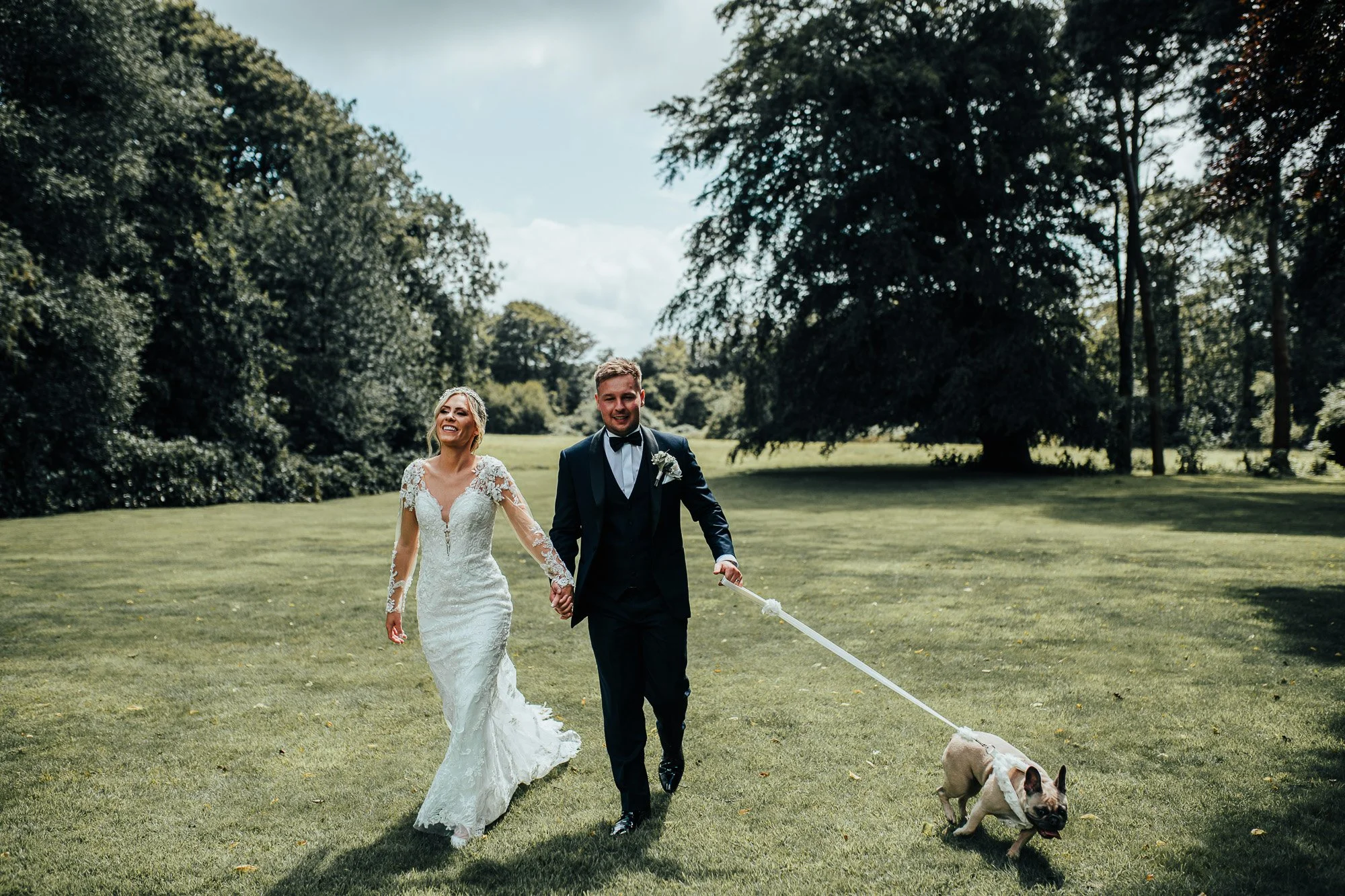 Bride and groom holding hands with a French Bulldog on a leash walking in a grassy area with trees.