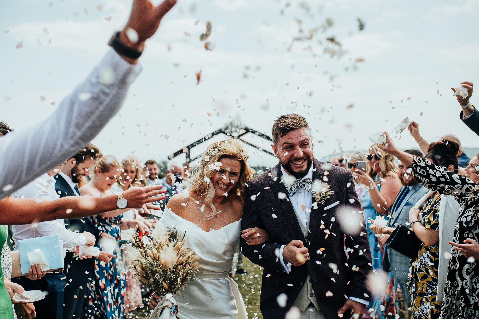 Bride and groom walking through confetti shower at an outdoor wedding, smiling with guests celebrating.