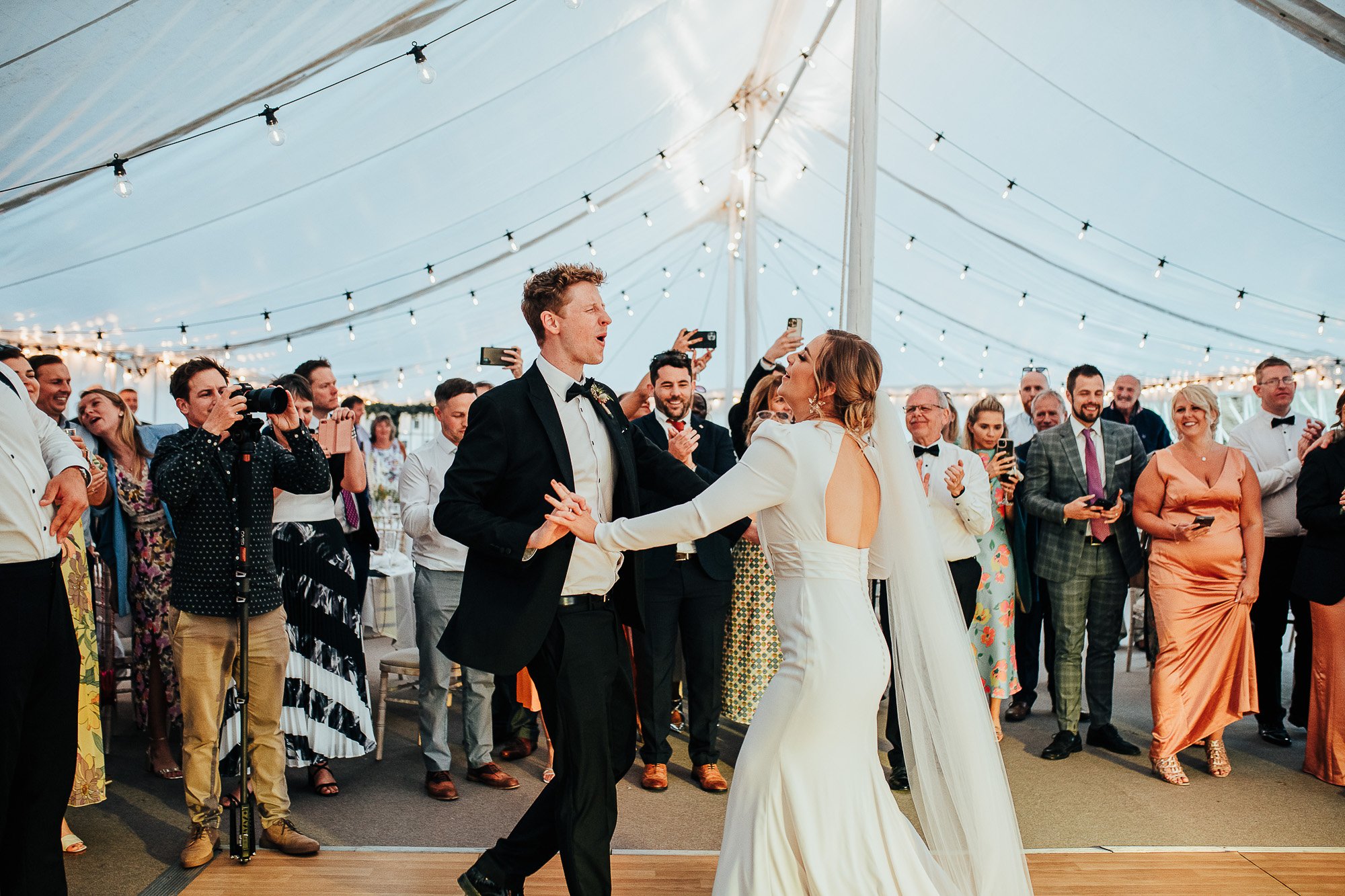 Bride and groom dancing under a tent with guests watching and taking photos at a wedding reception.