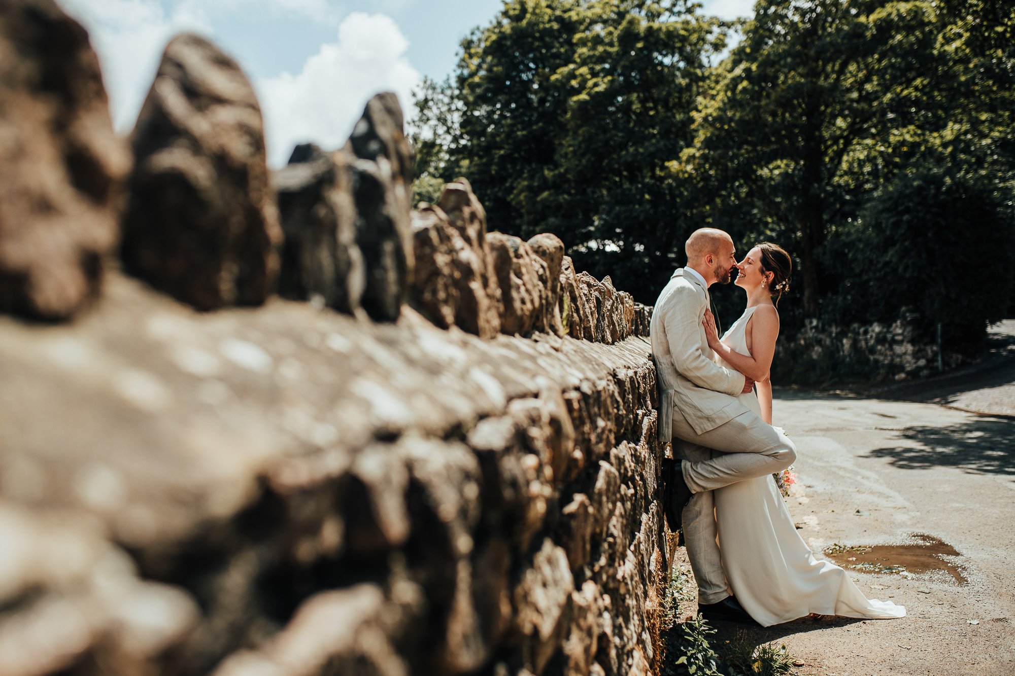 Bride and groom in wedding attire leaning against a rustic stone wall, smiling at each other, with trees and a road in the background.