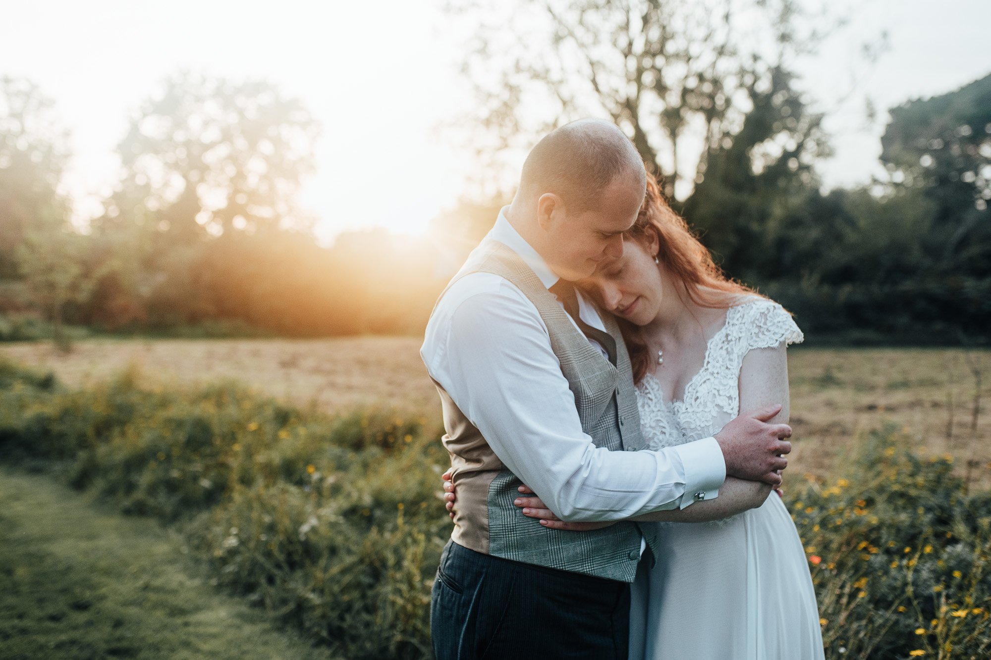 Bride and groom embracing outdoors at sunset, with a field in the background.