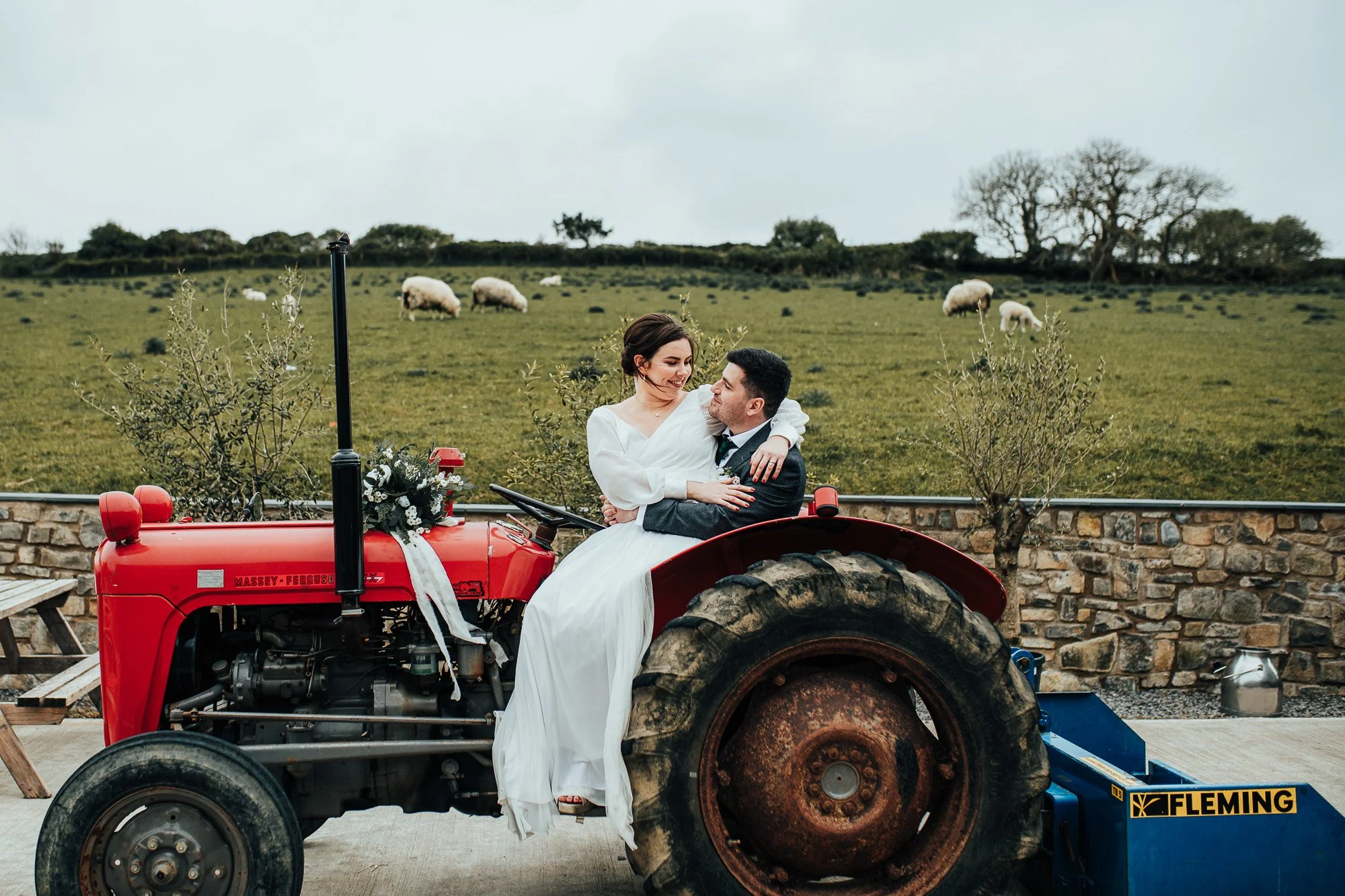 A bride and groom sitting on a red tractor in a rural setting with a field and sheep in the background.
