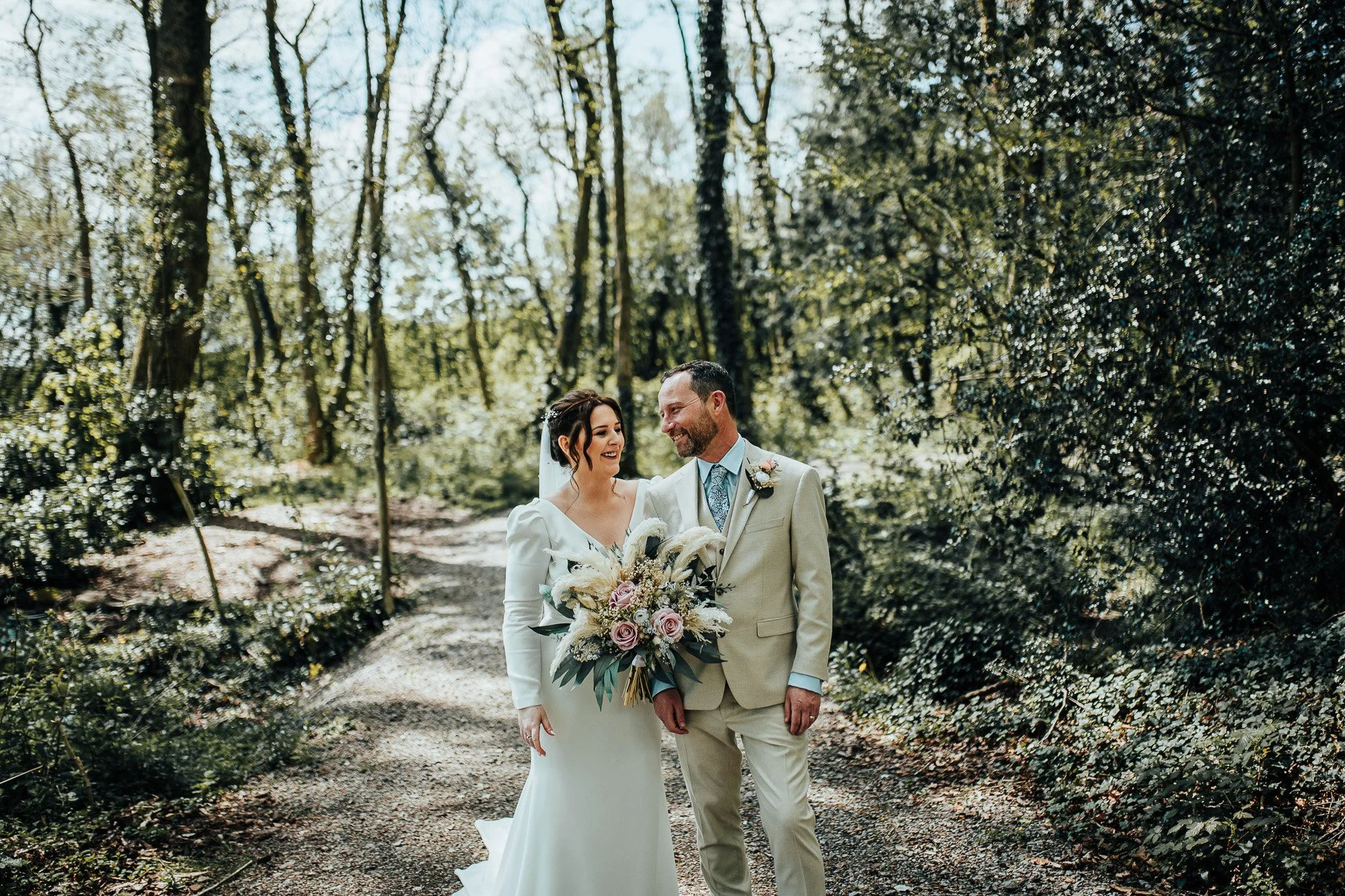Bride and groom smiling in forest, holding bouquet.