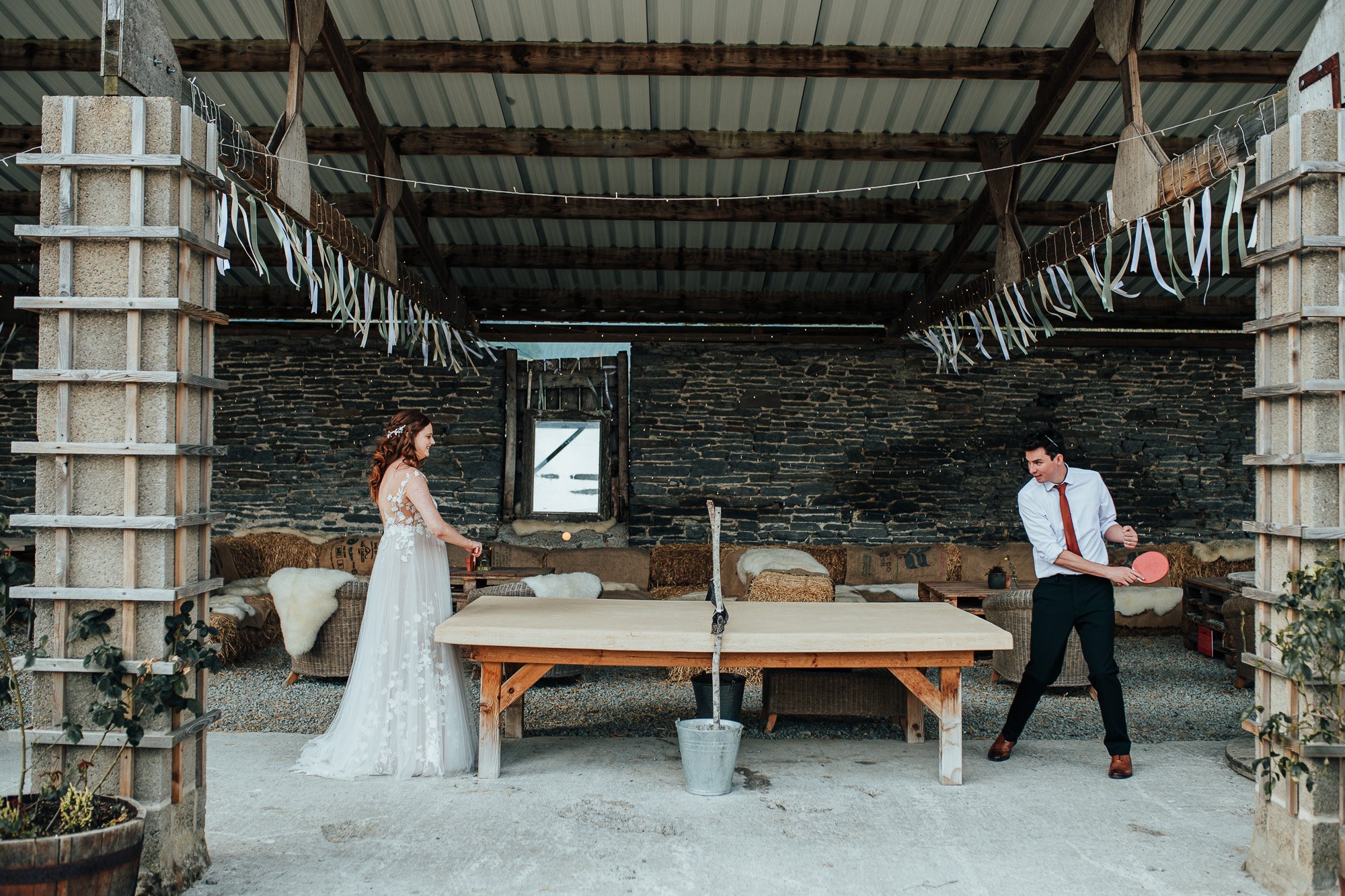 A bride and groom playing table tennis in a rustic barn setting. The bride is in a white wedding dress and the groom is in a white shirt and red tie. The table is makeshift, with a bucket holding the net. The barn has a stone wall and white streamers