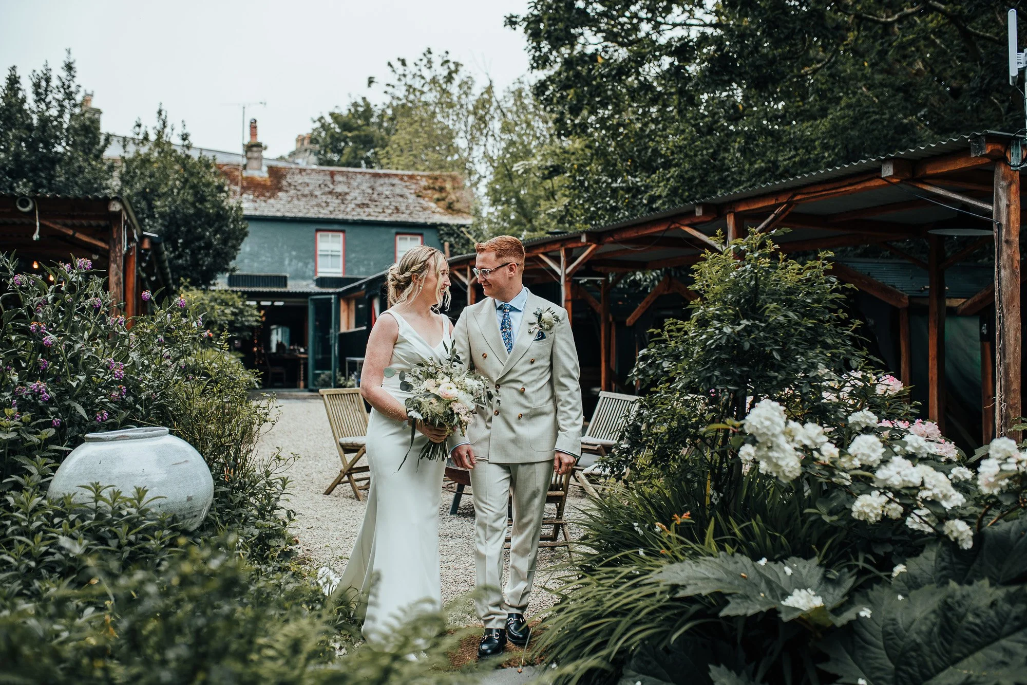 Bride and groom walking in garden, holding hands, with flowers and greenery around.