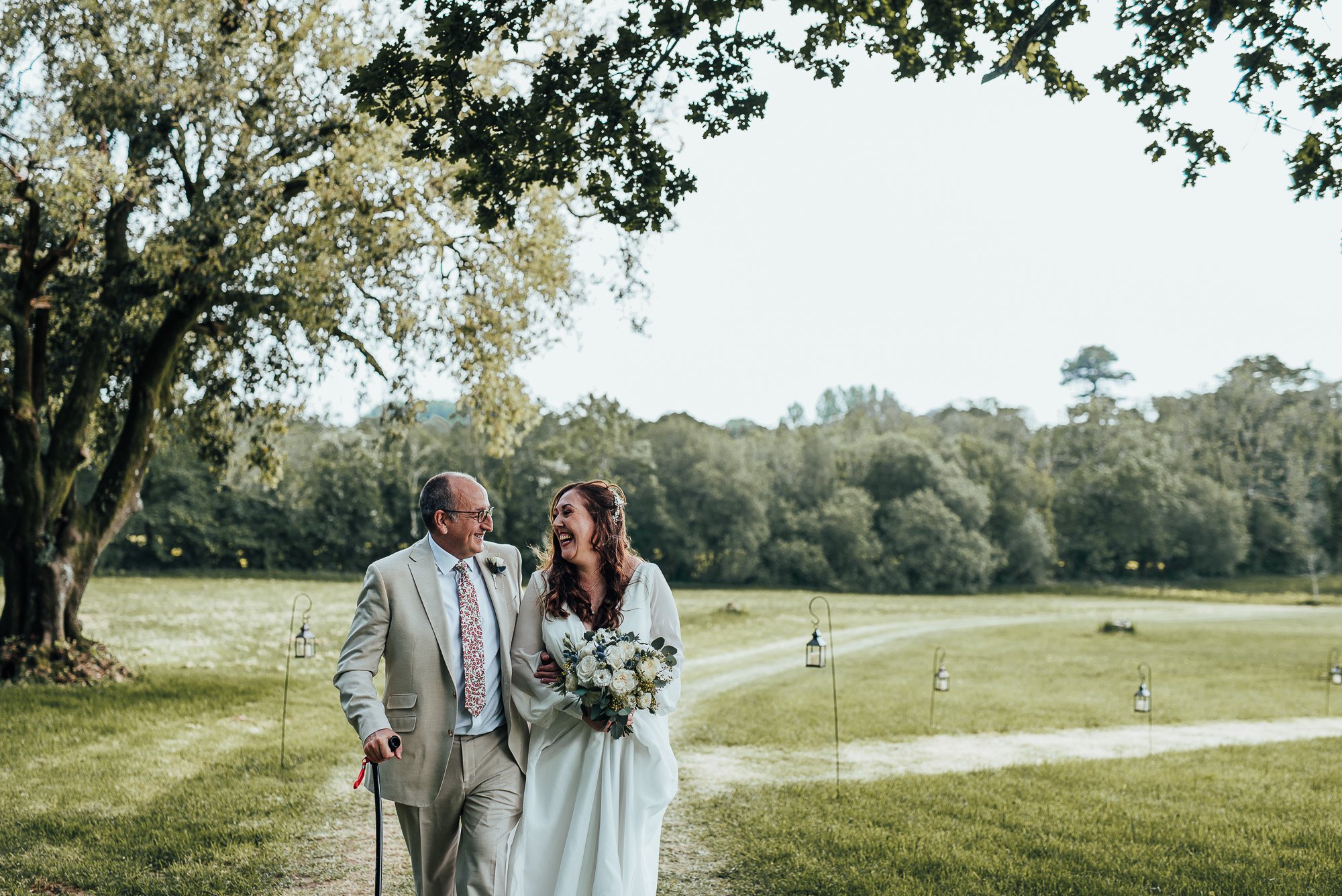 Bride and older man walking outdoors, smiling, with a green park-like background and hanging lanterns along the path, under tree branches.