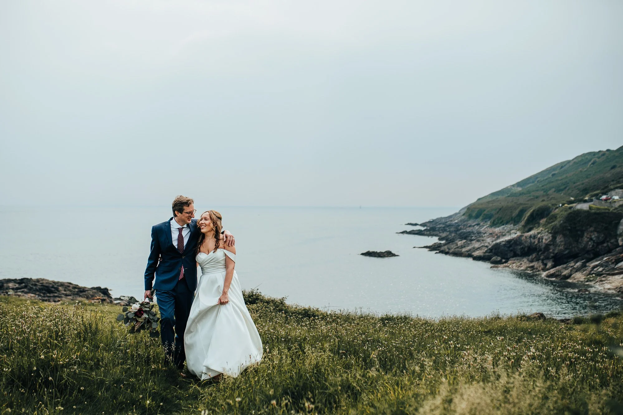 Bride and groom walking on grassy cliff by the sea.