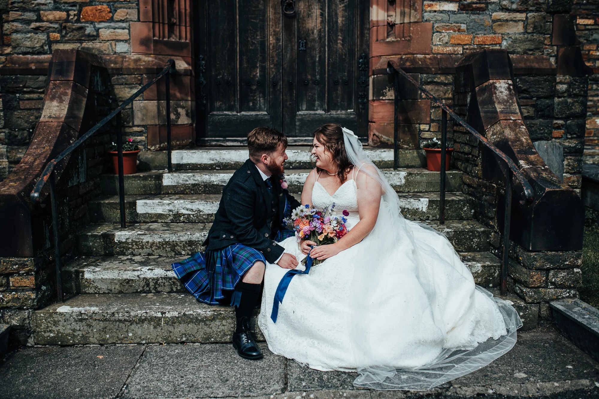 Bride and groom sitting on stone steps, smiling at each other. Bride in a white dress with a veil, holding a colorful bouquet. Groom wearing a traditional Scottish kilt and jacket. Background features stone building facade with dark wooden door.
