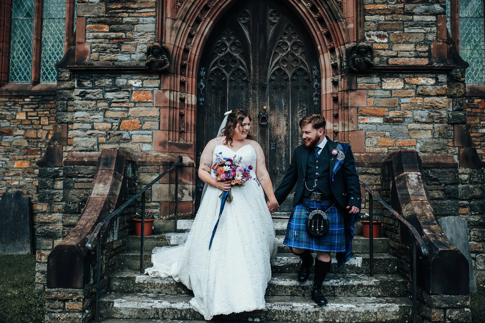 A bride in a white dress and groom in a kilt holding hands on stone steps, outside a historic church with a stone facade and arched door.