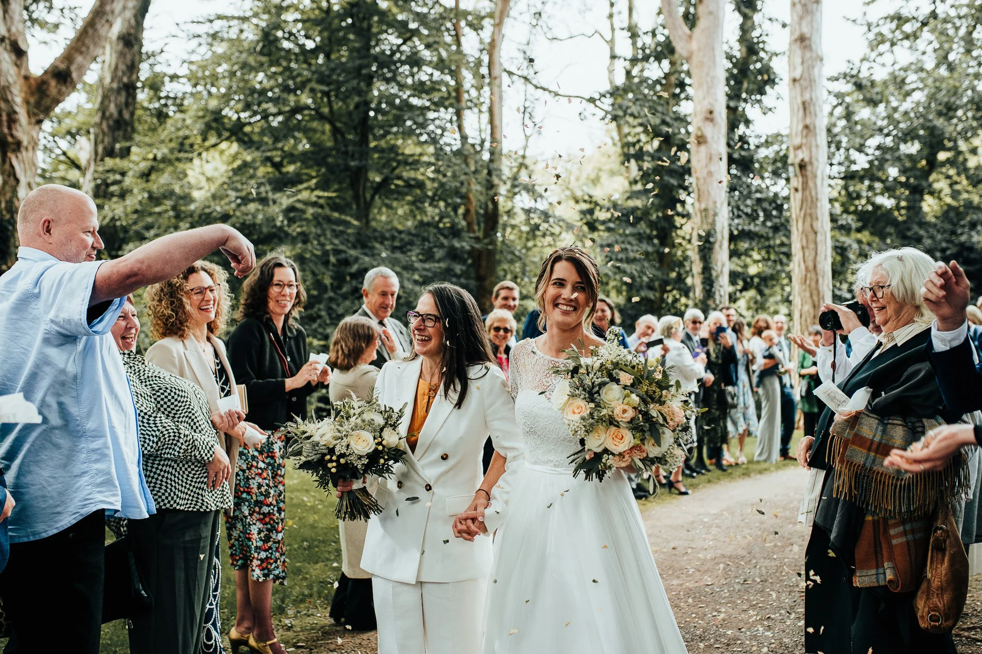 Two brides walking happily down an outdoor aisle surrounded by smiling guests at a wedding ceremony, with trees in the background.
