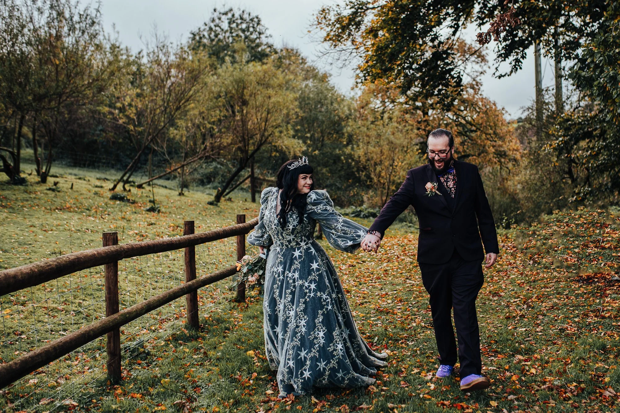 A couple walking hand in hand outdoors, woman in a blue star-patterned dress with a tiara, man in a dark suit, surrounded by autumn foliage.