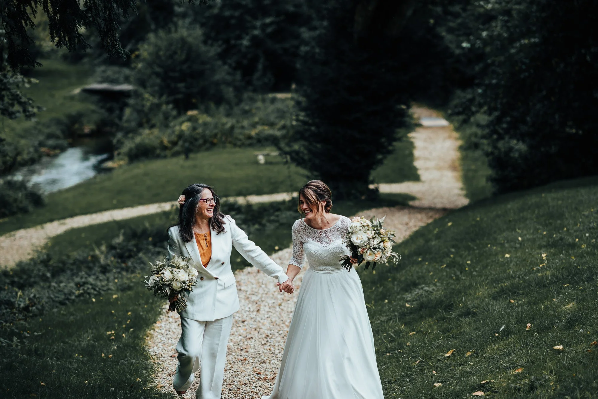 Two women holding hands, walking on a path in a garden. One is wearing a bridal gown, and the other is in a white suit, both holding bouquets and smiling.