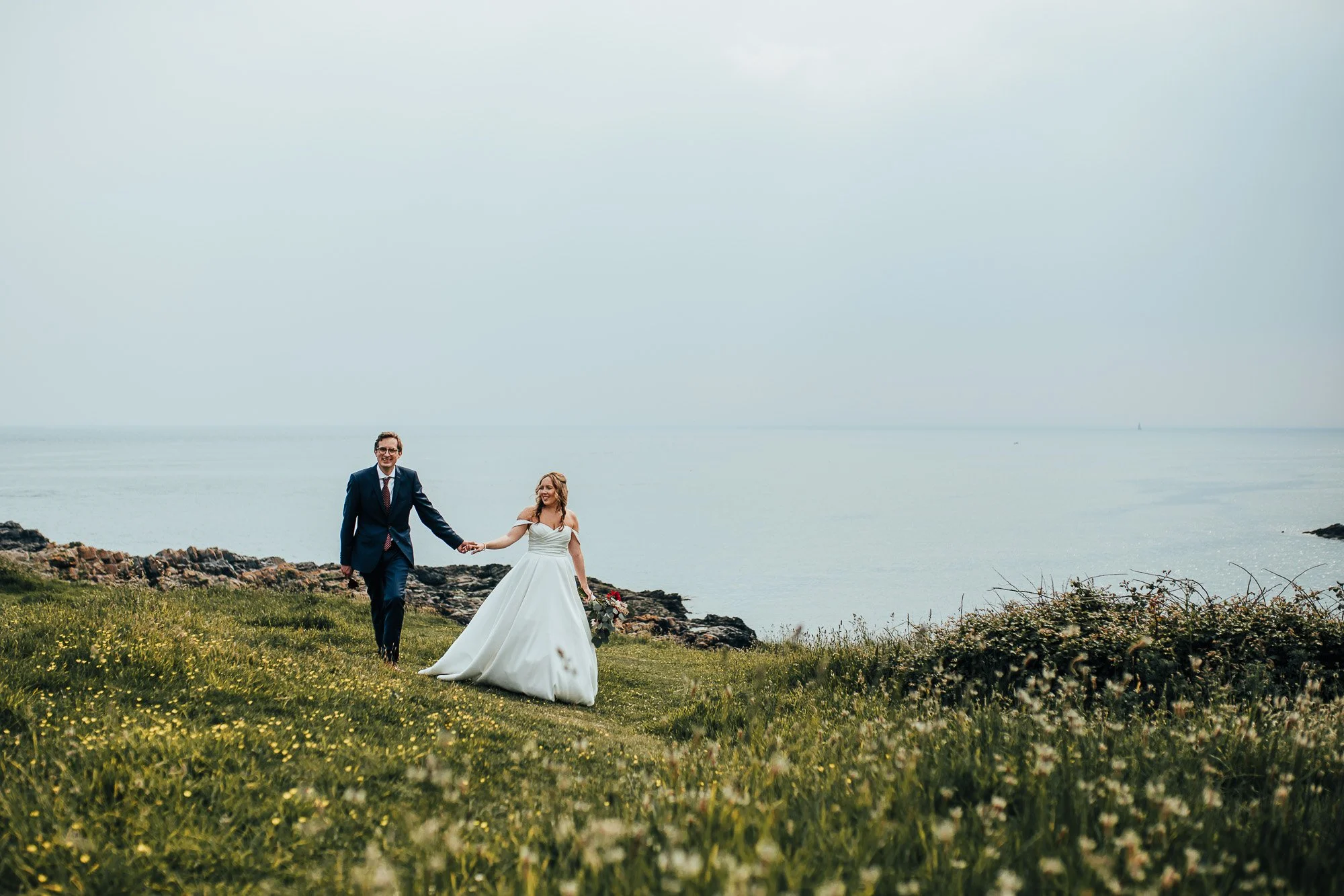 Bride and groom holding hands walking on grassy field near ocean.