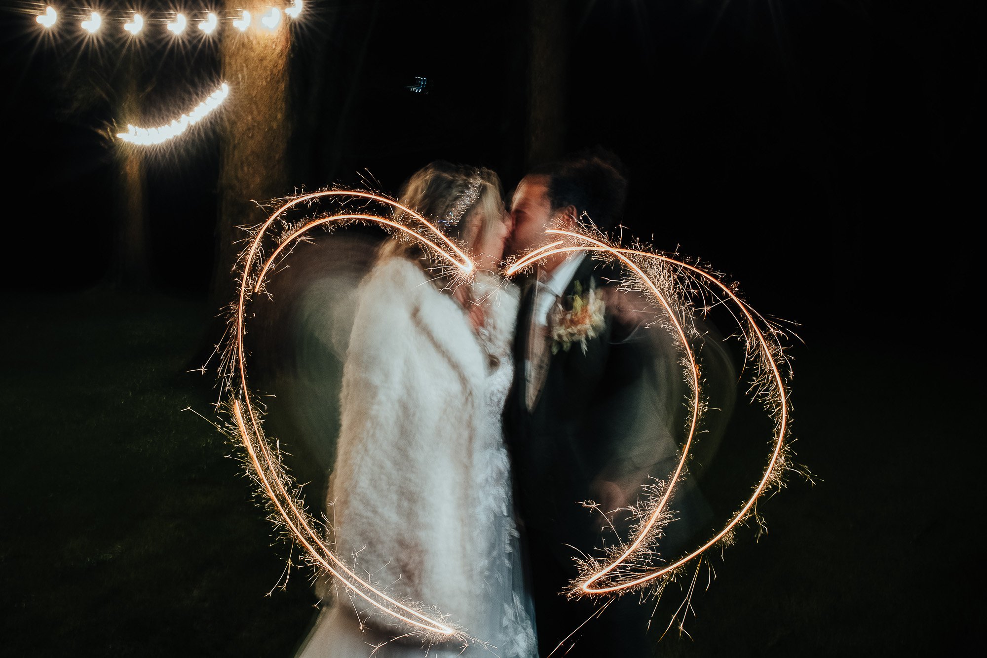 A couple kisses with sparklers creating a heart shape around them at night, with string lights in the background.