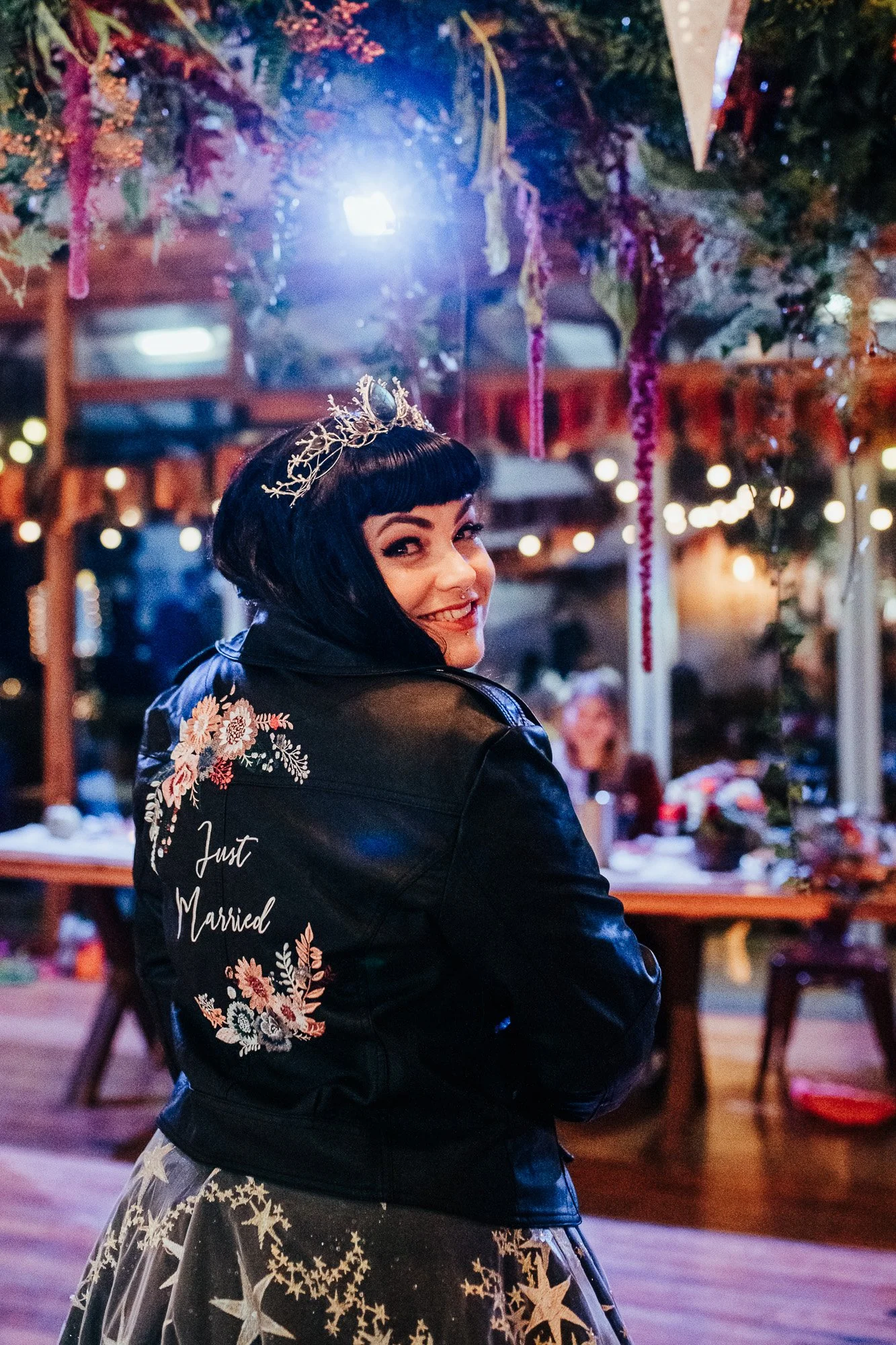 Bride wearing a decorated jacket with 'Just Married' embroidered, smiling in celebration, indoors at a wedding reception with floral decorations.