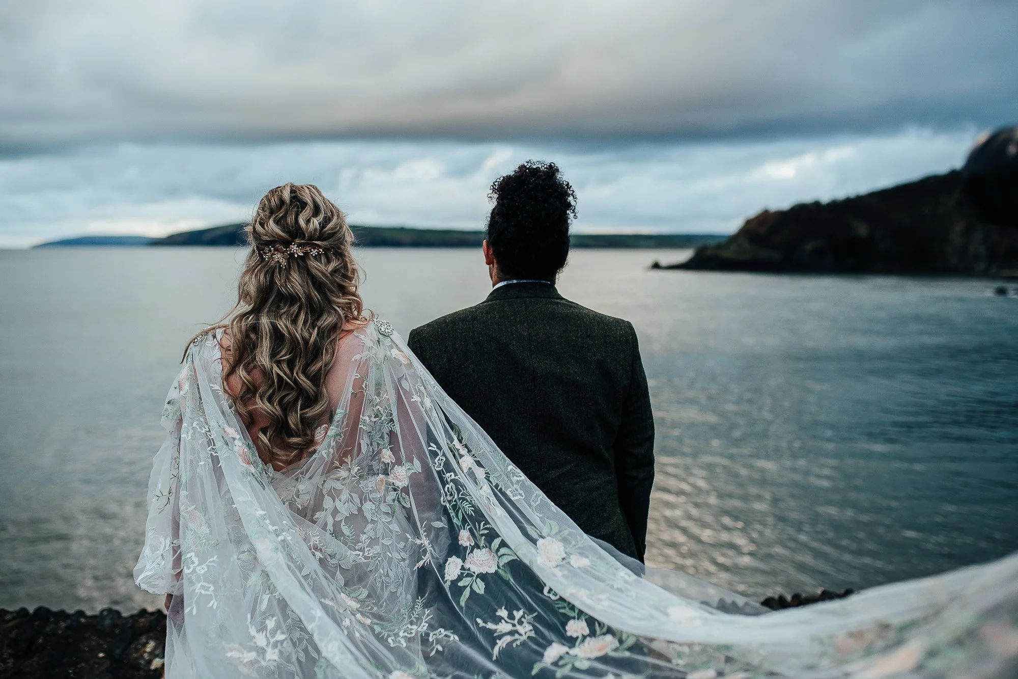 Bride and groom facing a scenic view of the ocean with a rocky coastline and cloudy sky.