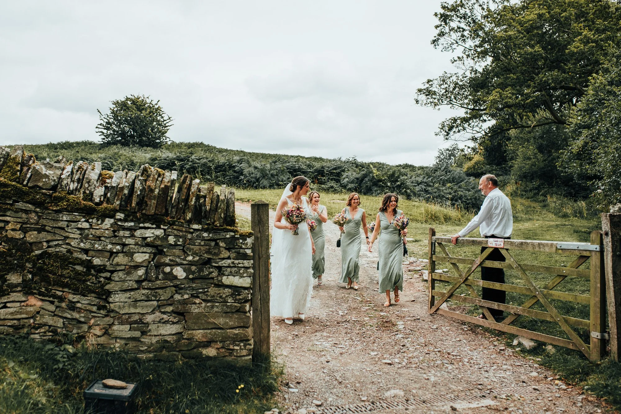Bride and bridesmaids walking through a rustic countryside gate with a stone wall and greenery, man holding the gate open.