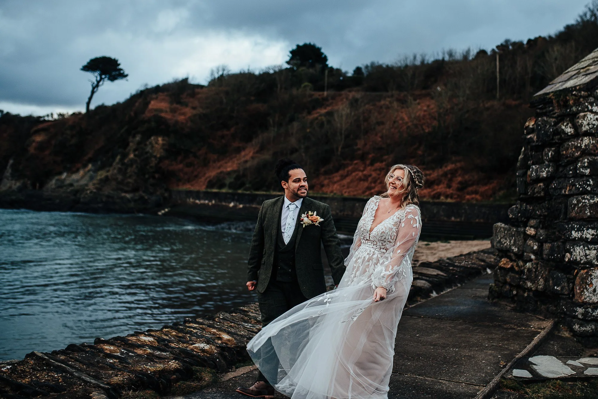 Bride and groom walking by a rocky shoreline, with the bride in a flowing dress and the groom in a dark suit, smiling against a backdrop of trees and a cloudy sky.