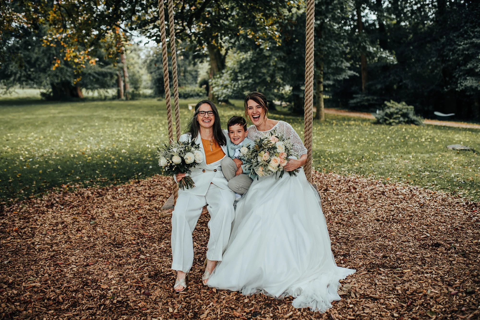 Two women in wedding attire and a child sitting on a swing with bouquets in a park setting.