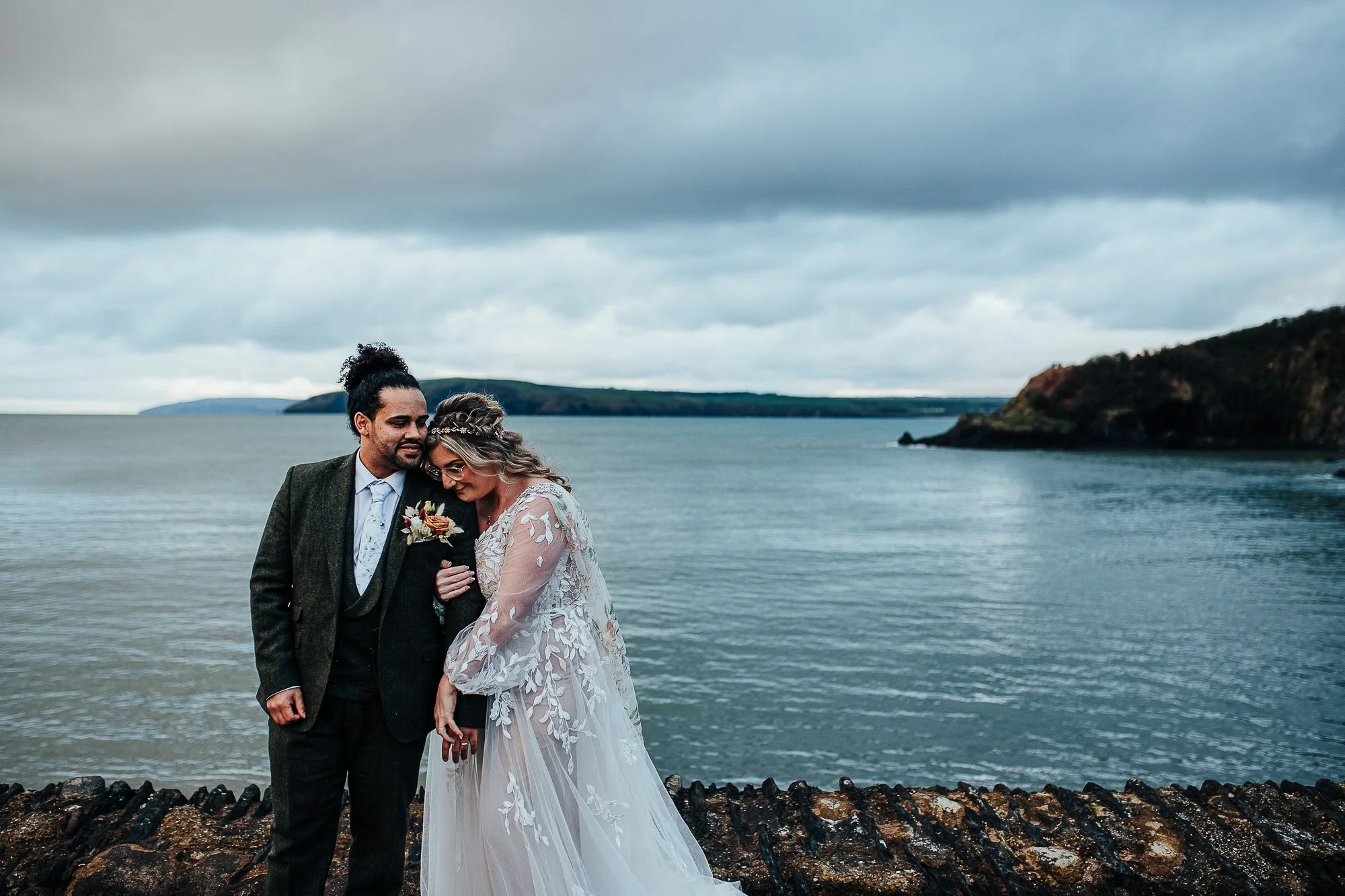 Bride and groom standing by the sea with a rocky coastline in the background.