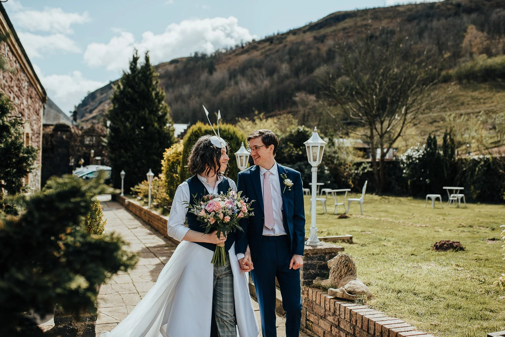 A couple holding hands outdoors, the woman holding a bouquet, both dressed semi-formally, with a garden and mountains in the background.