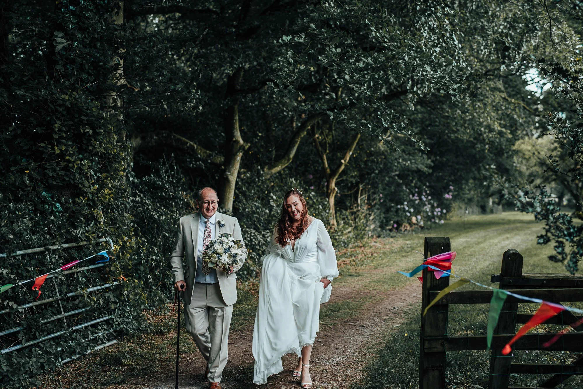 A bride in a white dress walks with an older man in a suit along a forest path, with colorful triangular flags on the sides. The man holds a bouquet and uses a cane.