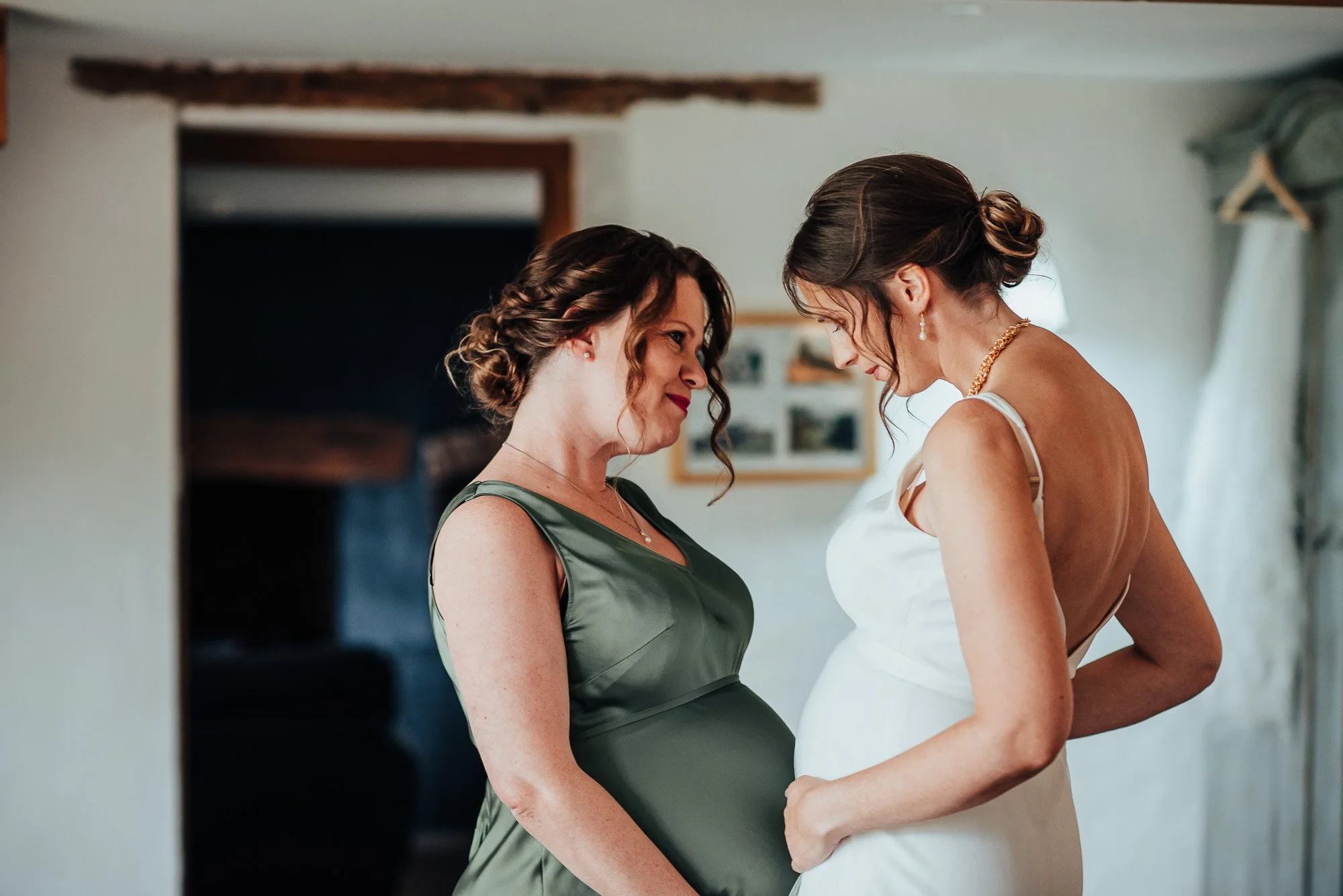 Two pregnant women in dresses happily facing each other in a cozy room.
