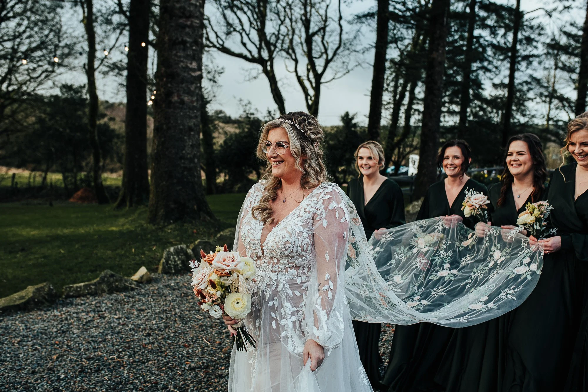 A bride in a floral lace wedding dress with a bouquet, surrounded by bridesmaids in dark dresses, outdoors with trees in the background.