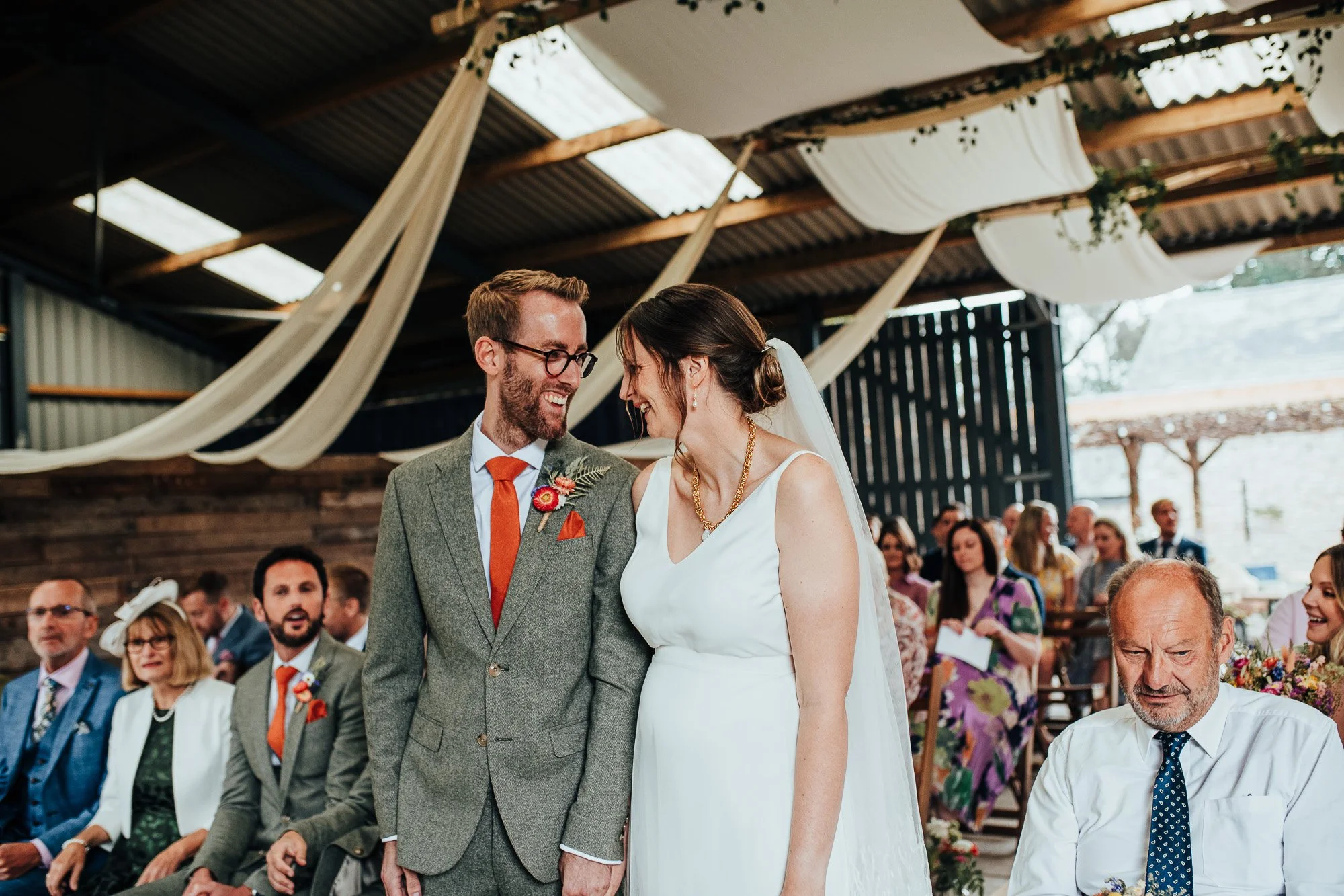 Bride and groom smiling at each other during their wedding ceremony in a decorated venue with guests seated.