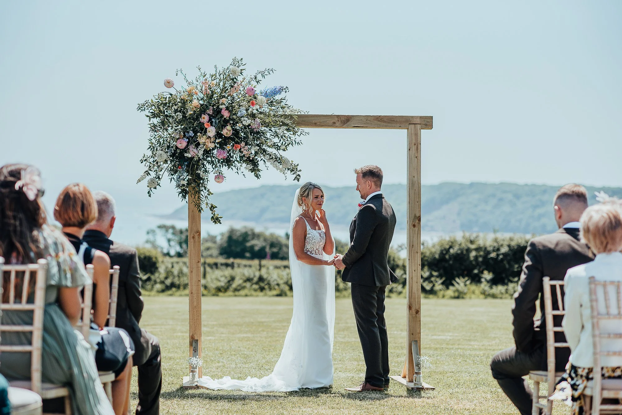 Outdoor wedding ceremony with bride and groom under a floral arch, surrounded by seated guests.