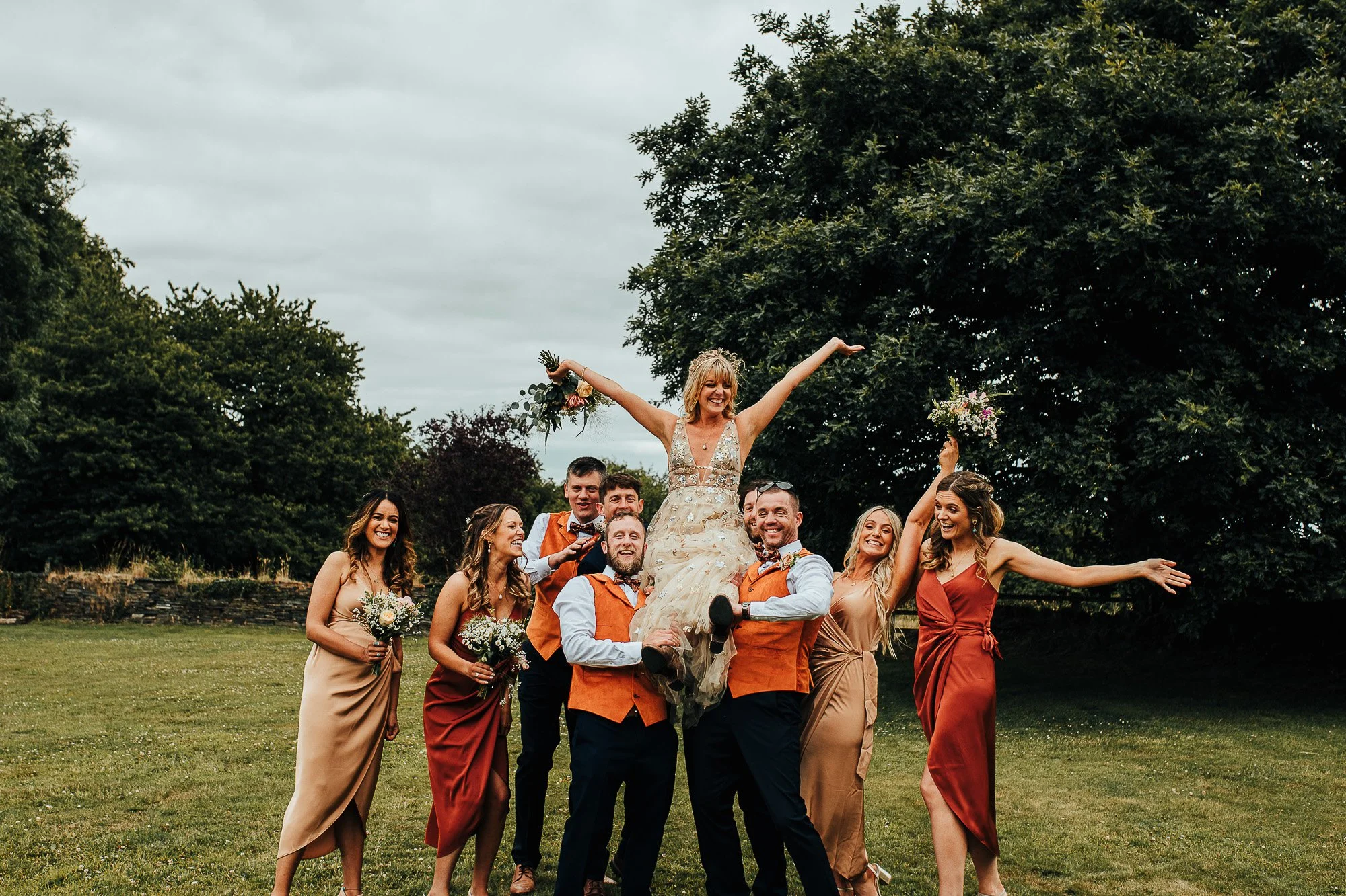 Bride lifted by groomsmen and bridesmaids in a garden setting, wearing formal attire, celebrating with joyful expressions.