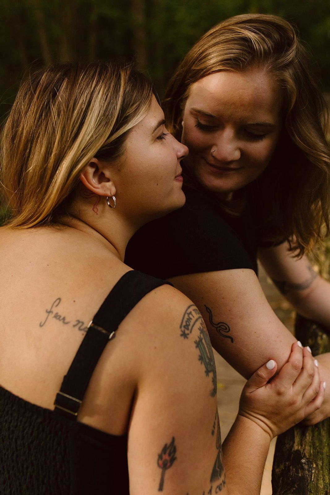 Two women sitting closely outdoors, facing each other, with their hands clasped. They have tattoos on their arms, and the background is blurred greenery.