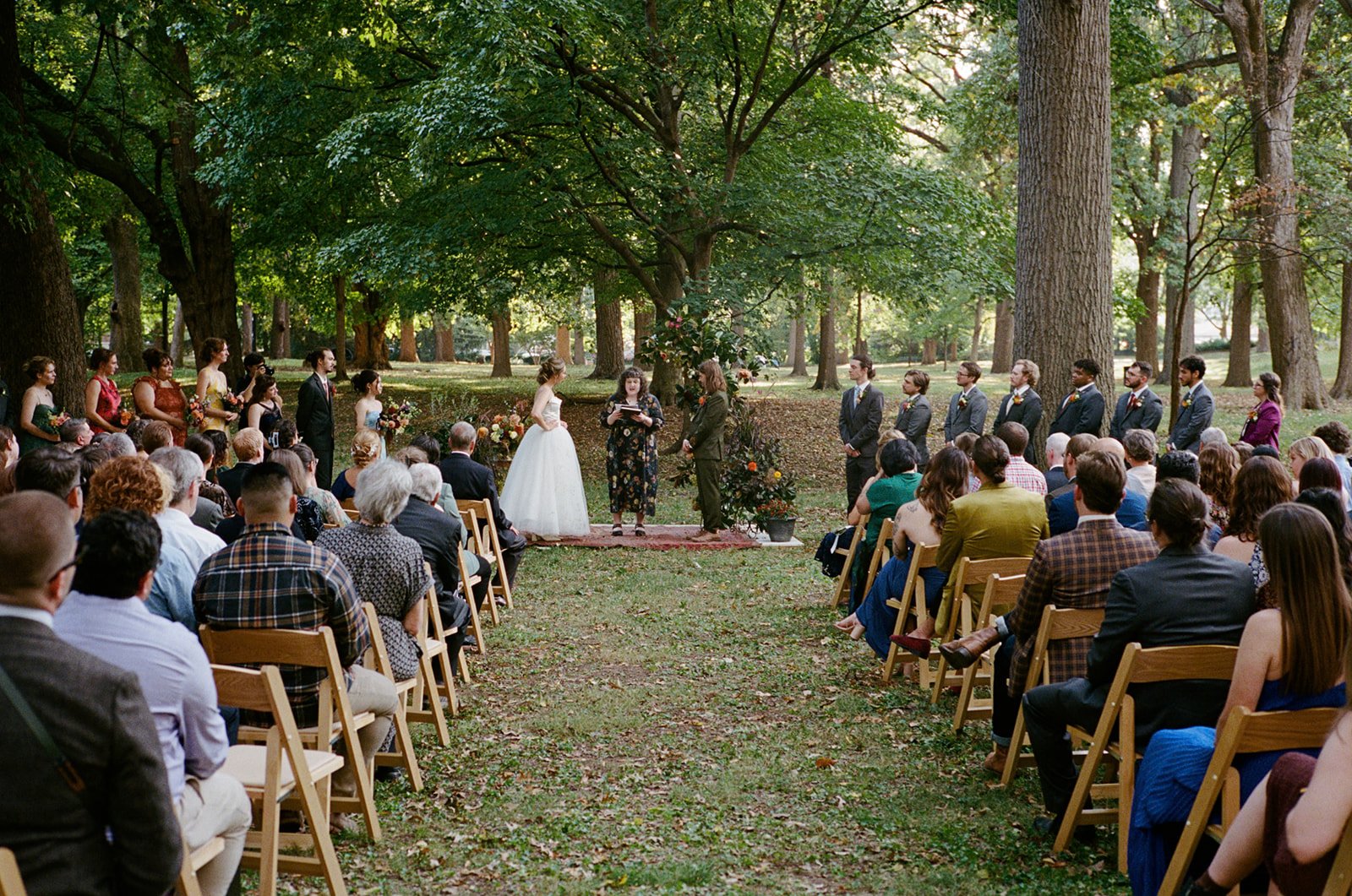 Outdoor wedding ceremony in a park with a bride in a white gown and groom in a dark suit facing officiant under large trees, guests seated on wooden chairs on either side.