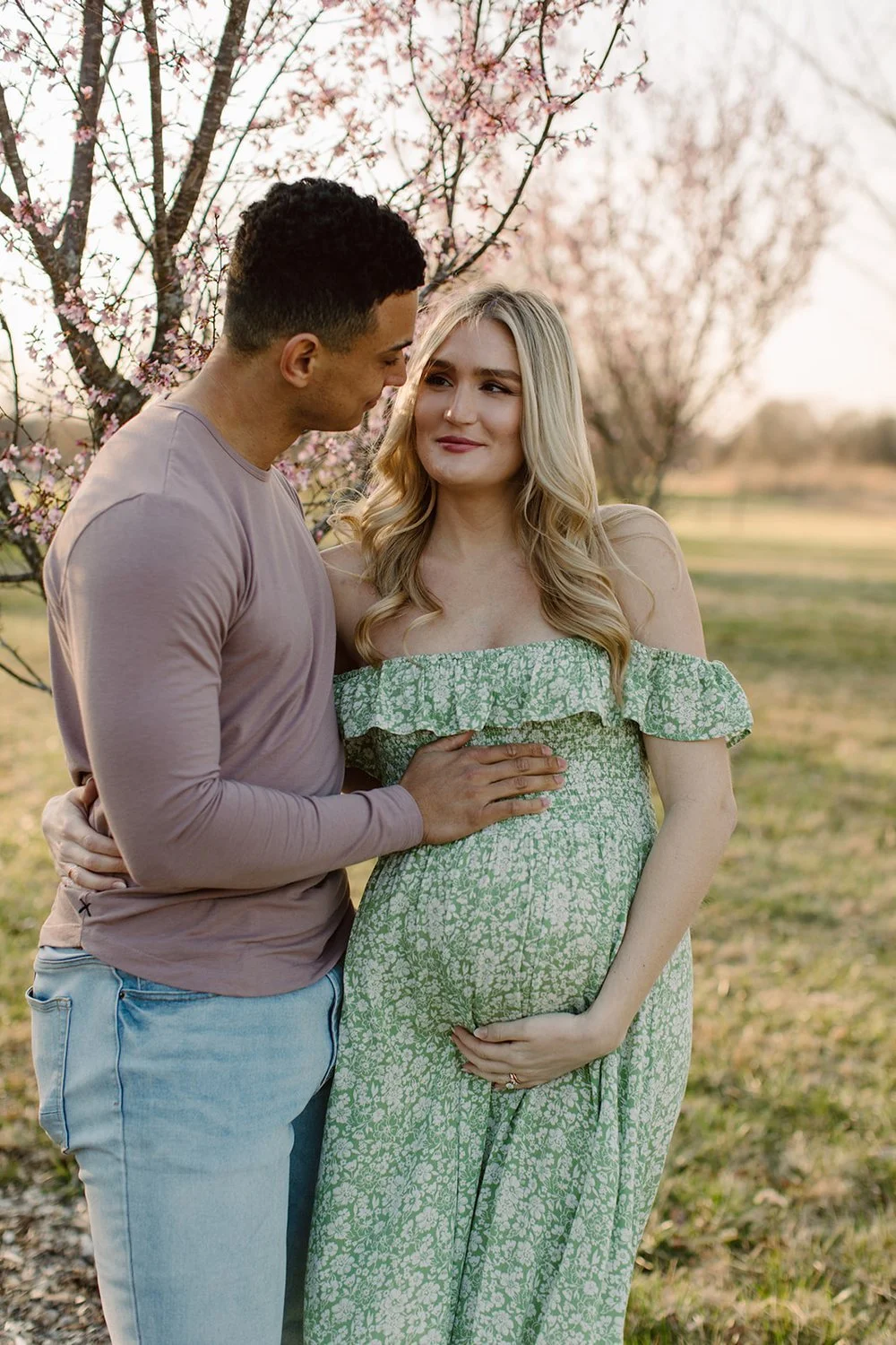 A pregnant woman and man standing outside near blossoming trees, with the woman holding her belly and the man touching her arm, during sunset.