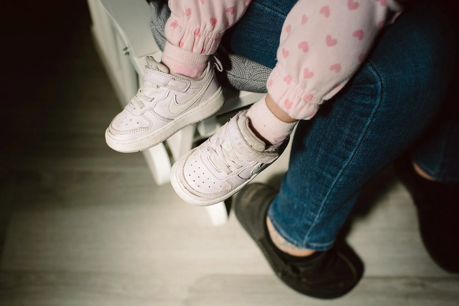 A child wearing pink and white sneakers, pink socks, pink pants with hearts, sitting on an adult's lap. The adult is wearing blue jeans and black shoes.