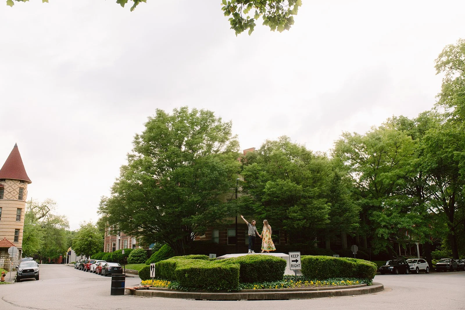 A couple standing on a small podium in a circular green space with bushes, trees, and flowers, holding hands with one arm raised, in a small town or neighborhood street.