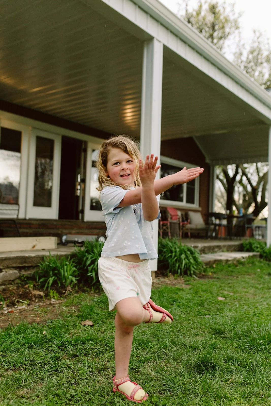 A young girl with curly blonde hair wearing a light blue shirt, white shorts, and pink sandals playing outside on the grass in front of a house with a porch.