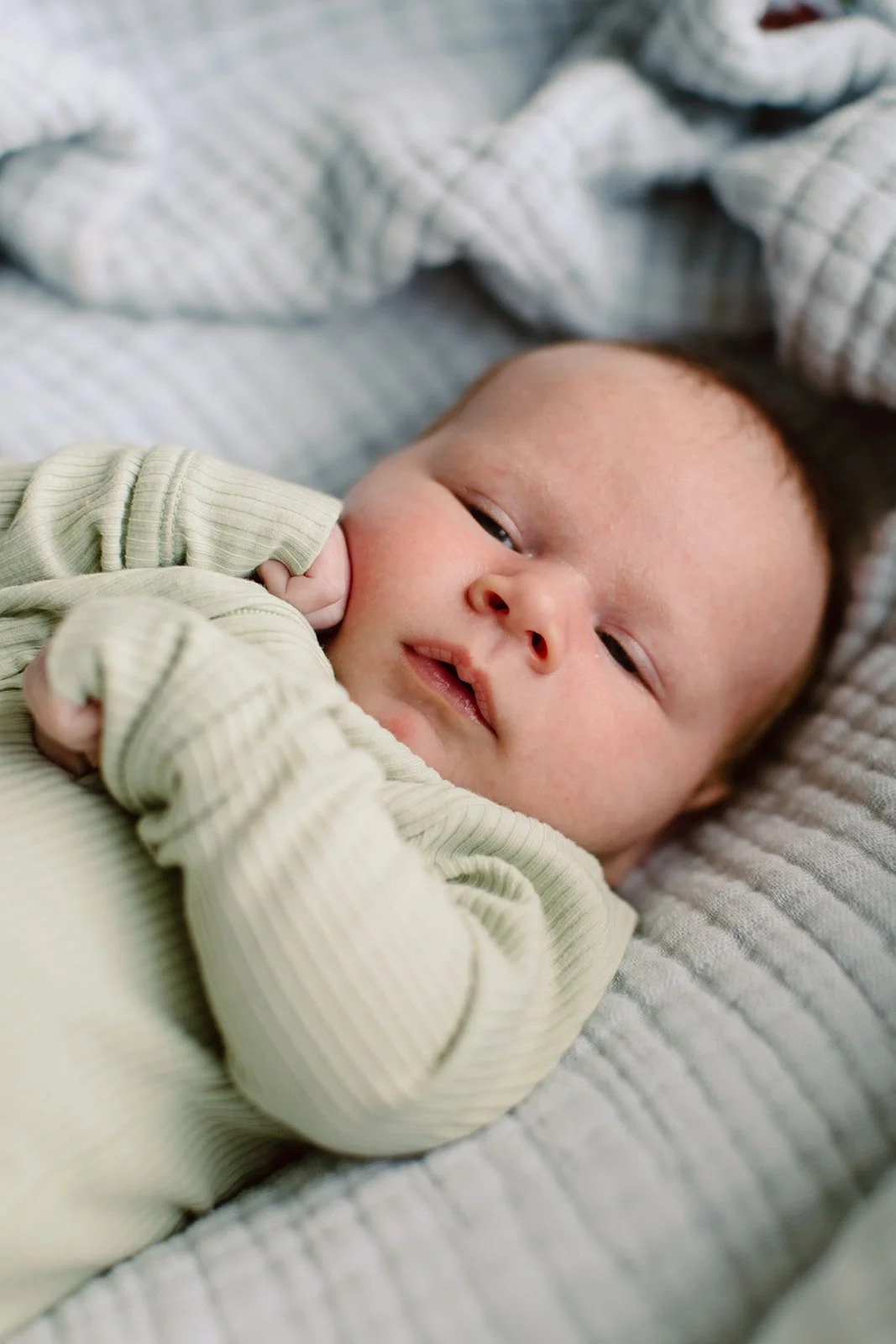 A baby lying on a bed, wearing a light green ribbed long-sleeve shirt, looking at the camera with relaxed eyes, and resting its head on a gray textured blanket.
