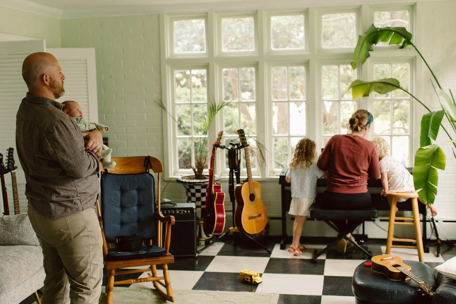 A man holding a baby watching a woman and two children playing a piano near large windows in a sunlit room with musical instruments, plants, and checkered flooring.