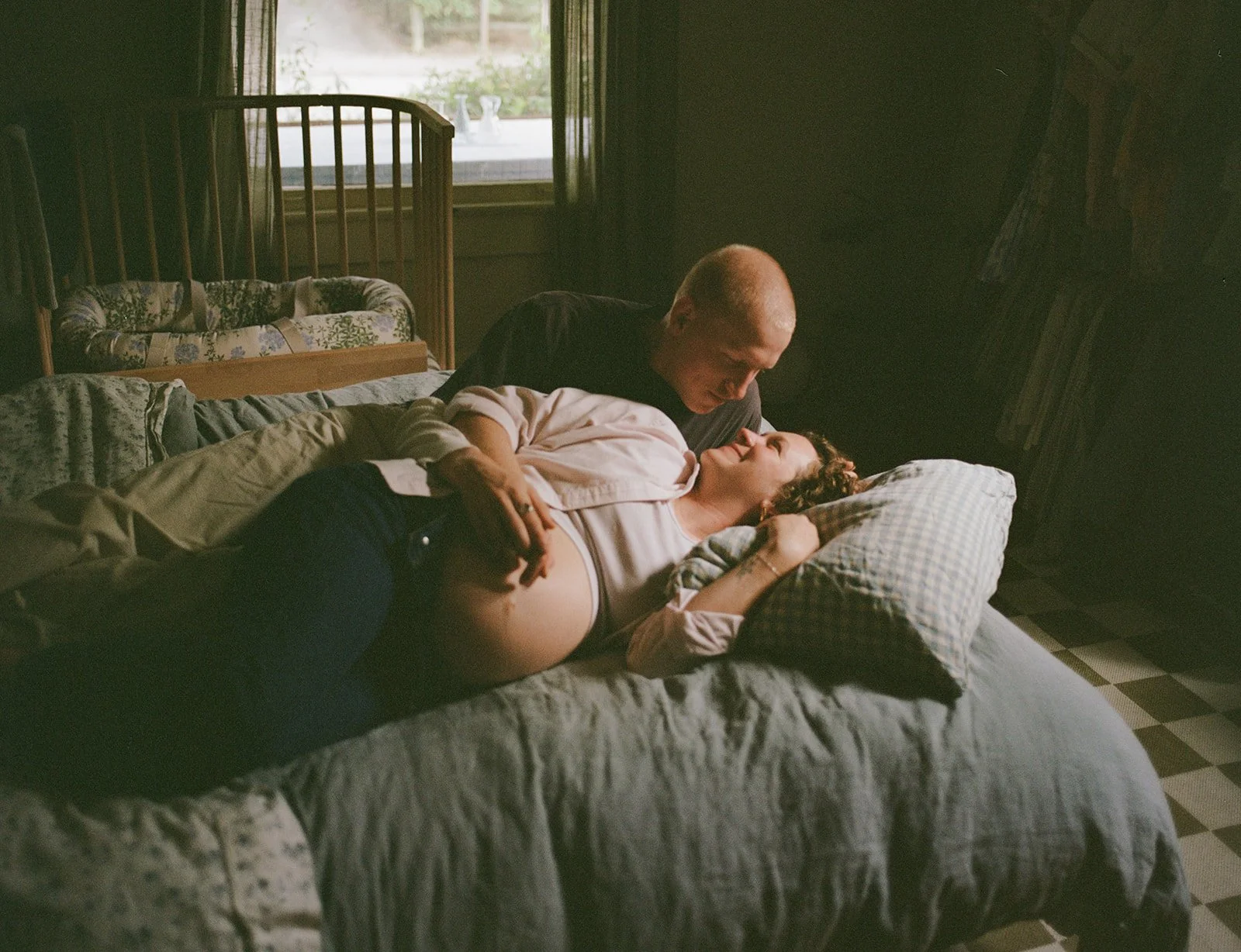 A couple lying on a bed in a bedroom, with sunlight coming through the window behind them. The man is leaning over the woman, and they are looking at each other fondly.