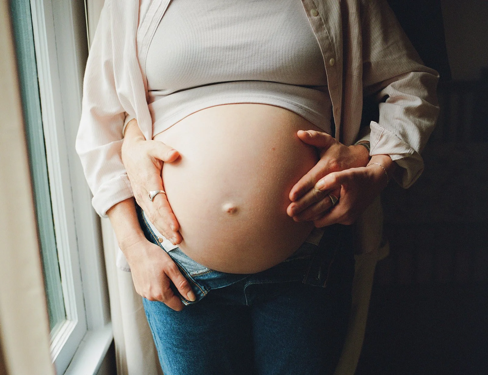 A pregnant woman gently cradles her baby bump with both hands while standing near a window.