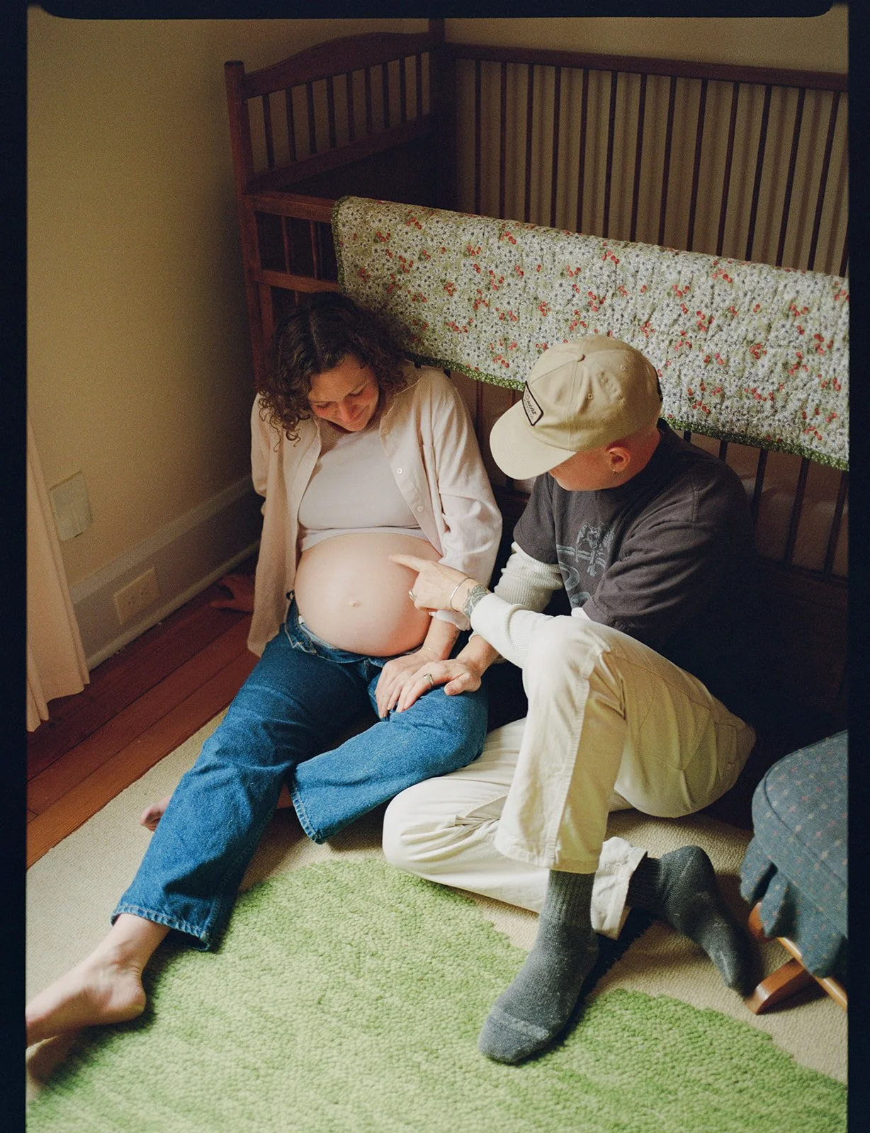 Pregnant woman sitting on the floor with a man pointing at her belly, both smiling. Bed with a floral blanket behind them.
