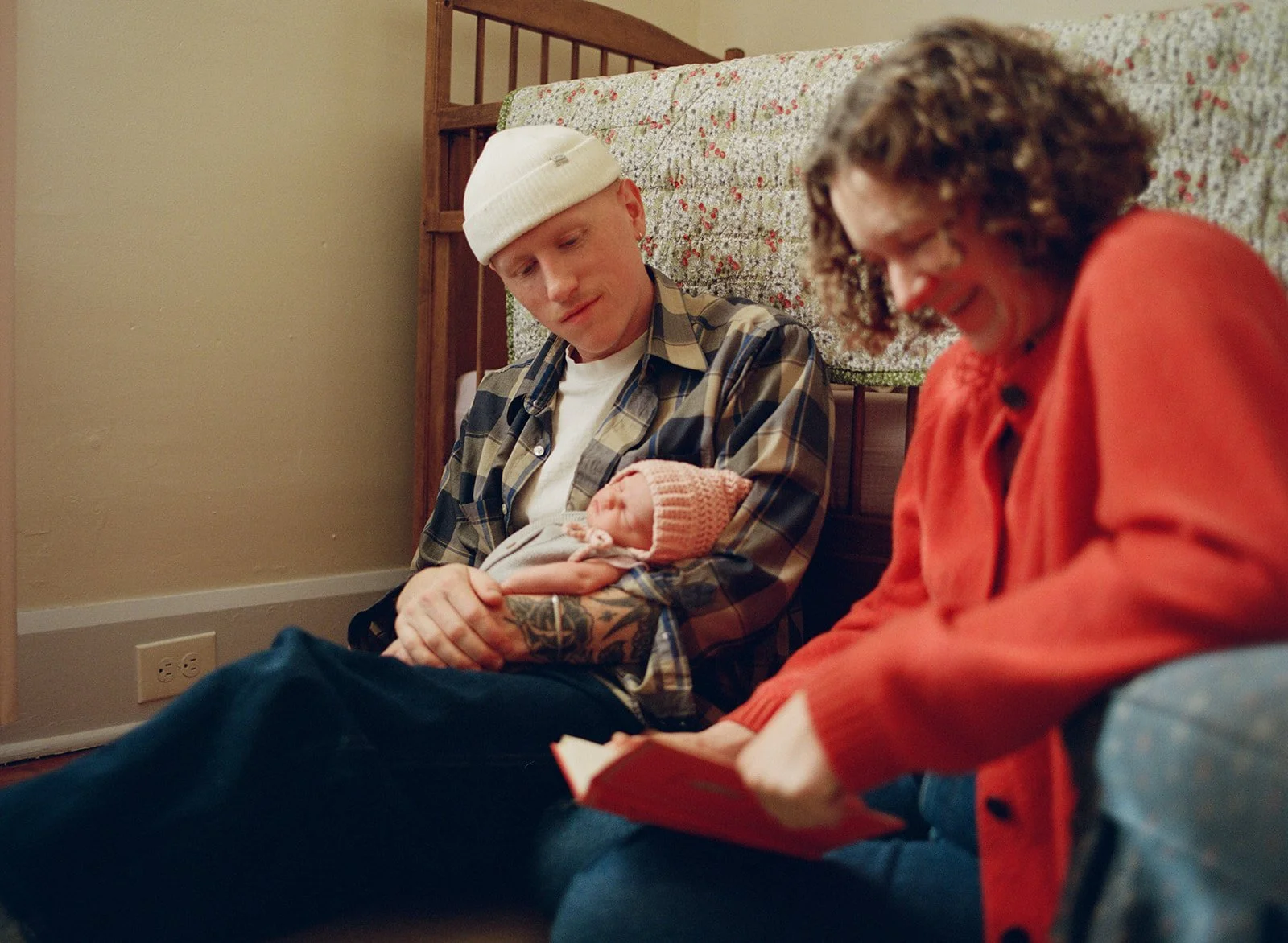 A young man with a white beanie and plaid shirt sitting on the floor, holding a newborn baby wrapped in a blanket, with a woman in an orange sweater sitting next to him looking at a book on her lap, in a bedroom with a bed and floral bedspread in the background.