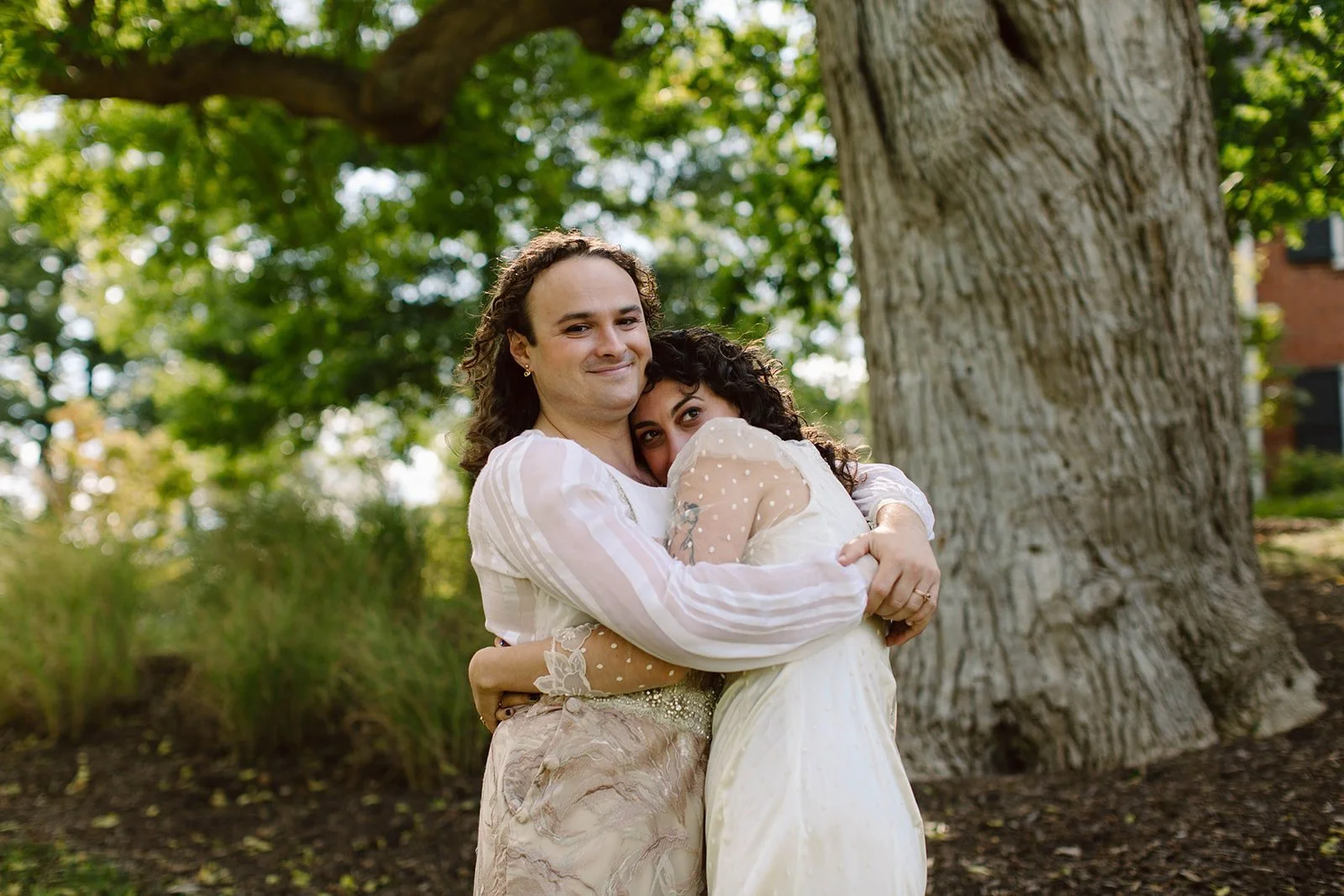 Two women embracing outdoors in front of a large tree. One woman is smiling while the other is resting her head on the first woman's shoulder. They are both wearing light-colored clothing.
