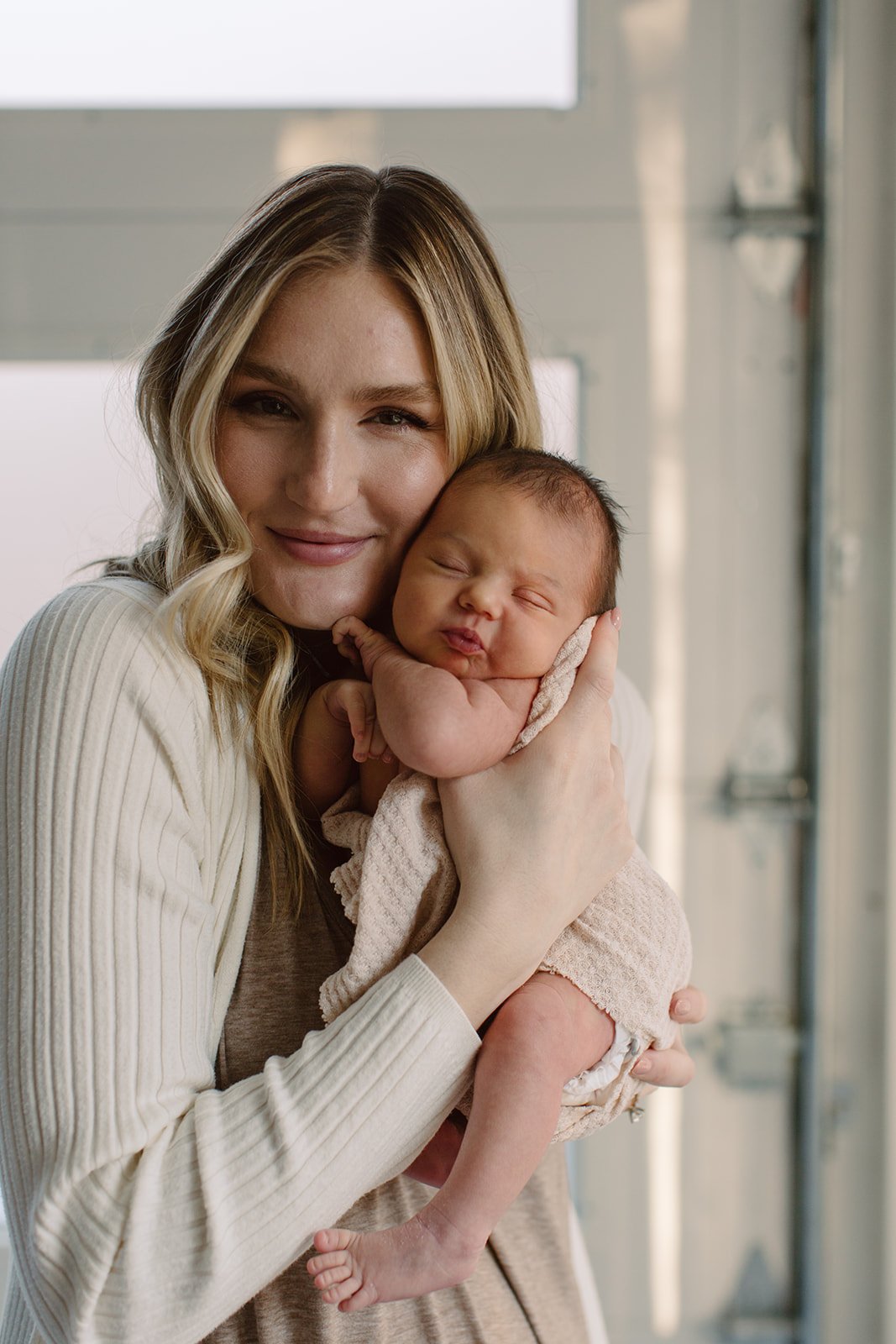 A woman with blonde hair holding a sleeping newborn baby close to her face, indoors near a large window.