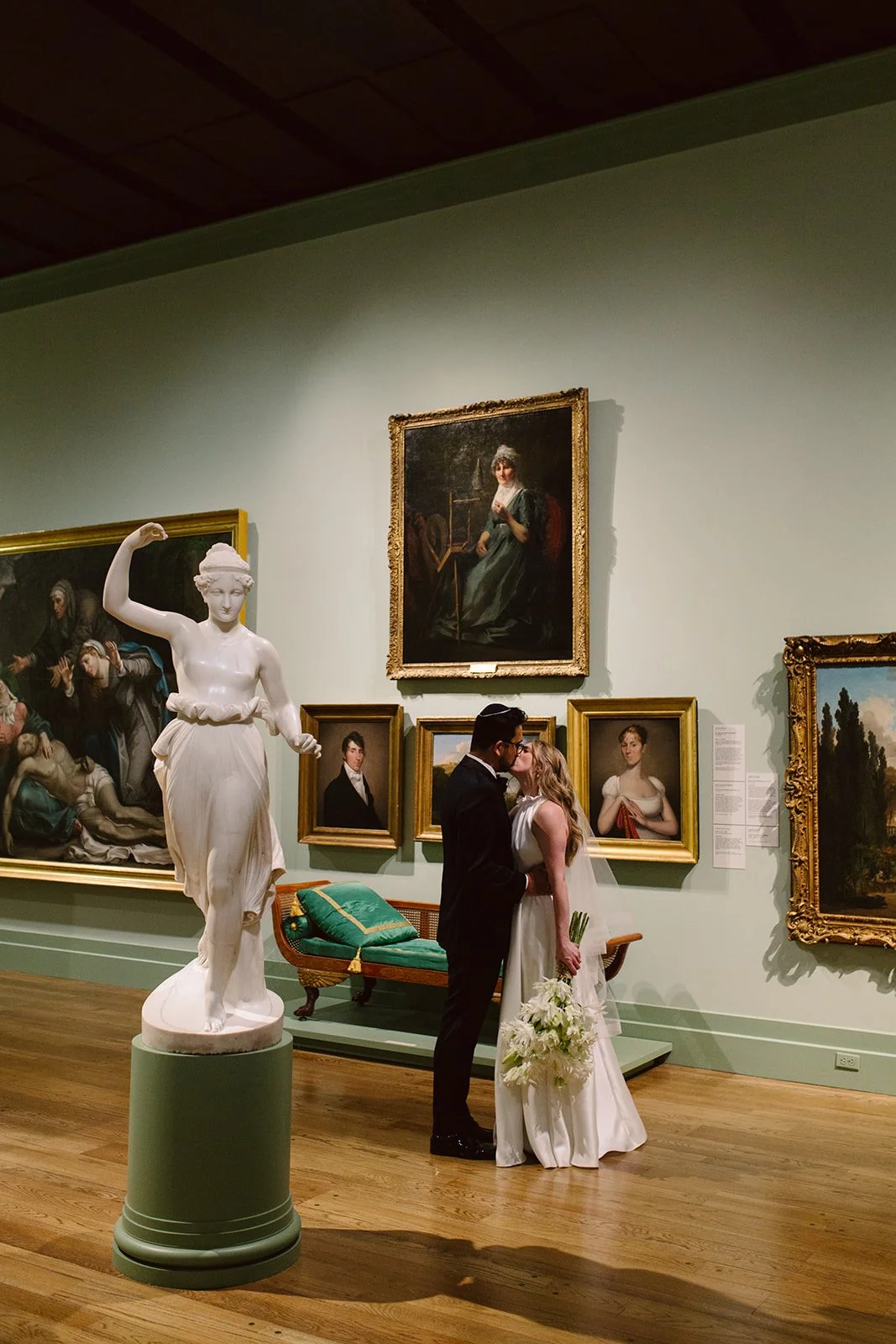 A newly married couple kissing in an art museum, surrounded by classical paintings and sculptures.