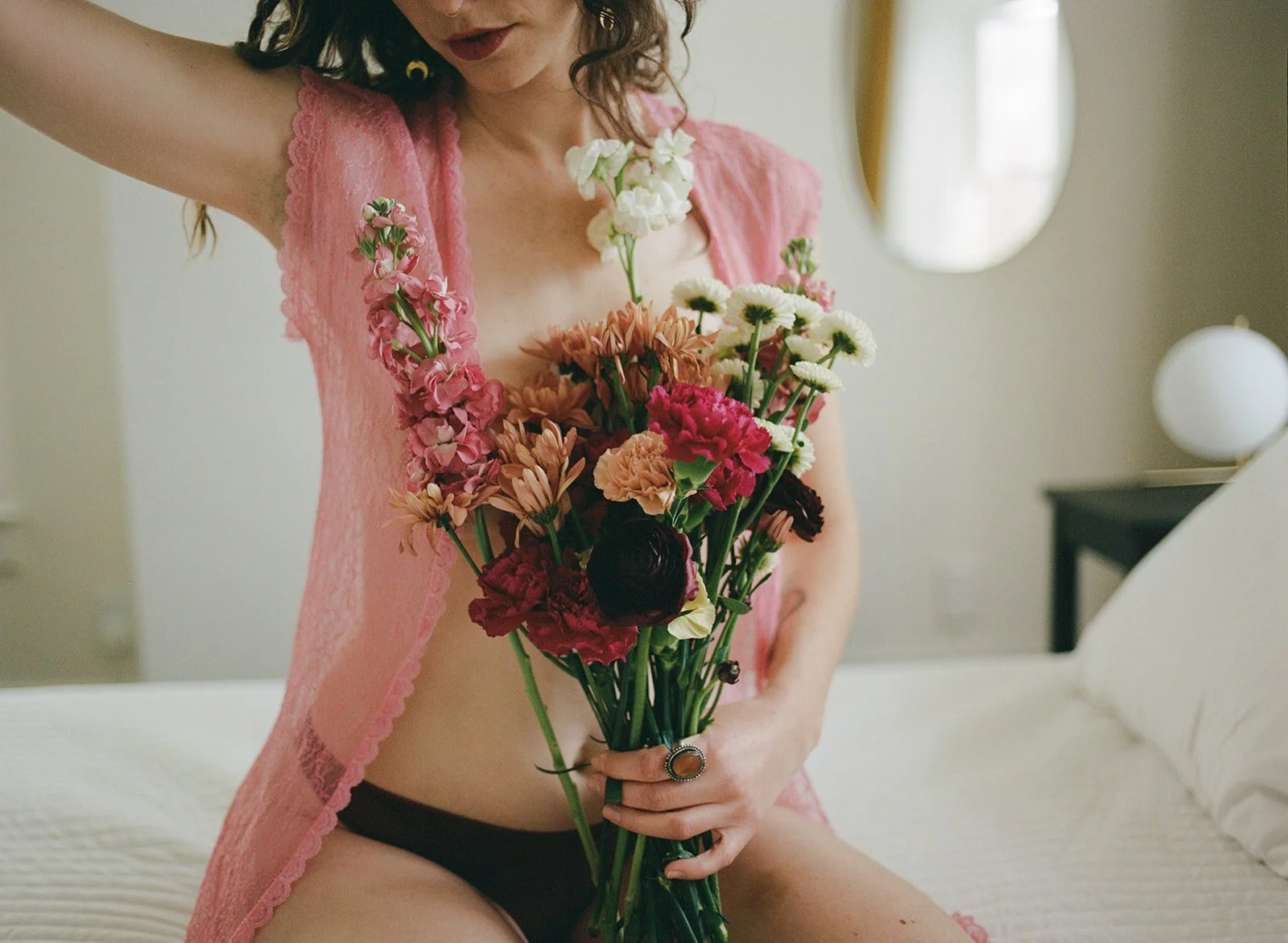 A woman wearing a pink lace robe and black underwear is sitting on a bed holding a large bouquet of colorful flowers, with a mirror and bedside lamp in the background.