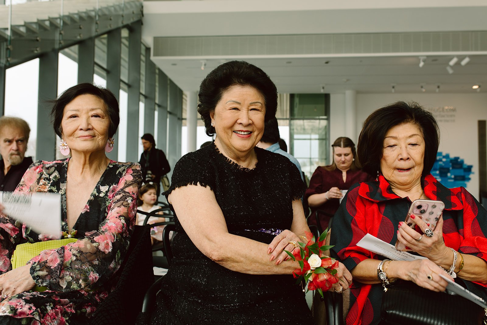 Three elderly women sitting indoors at an event, with the woman in the middle smiling and holding flowers, while the woman on the right looks at her phone, and the woman on the left looks forward. Other people are visible in the background.