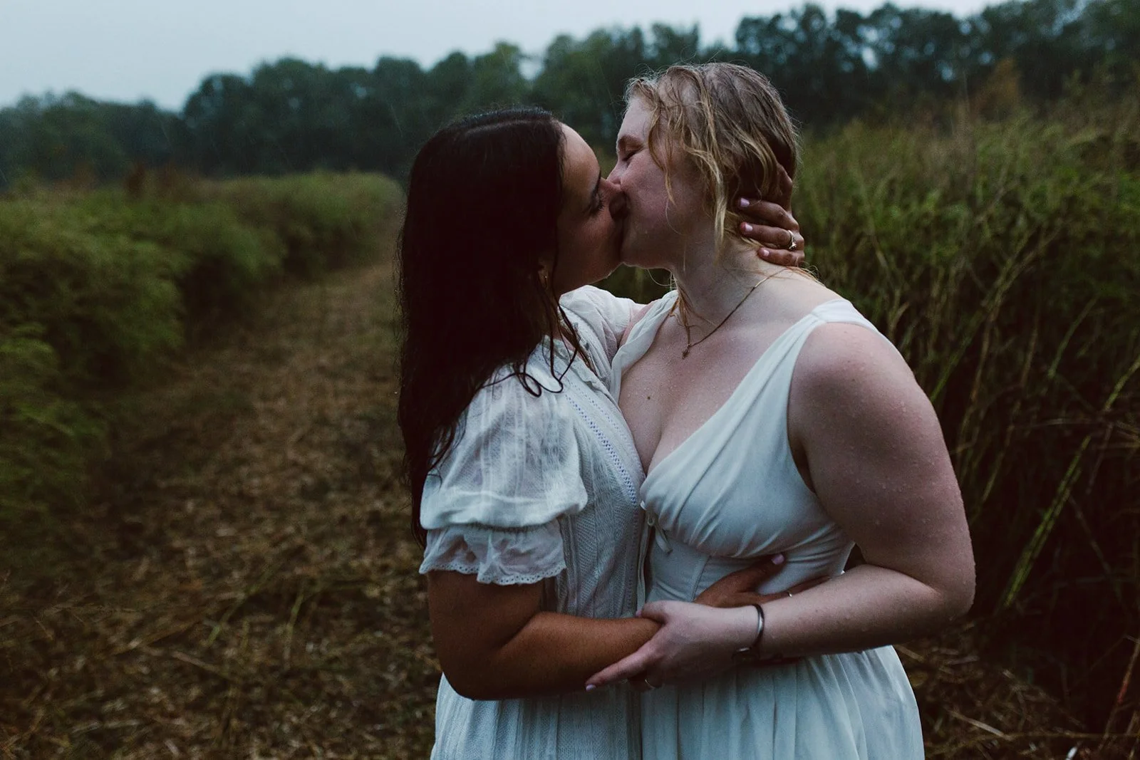 Two women in white dresses sharing a kiss in a natural outdoor setting during what appears to be rain.